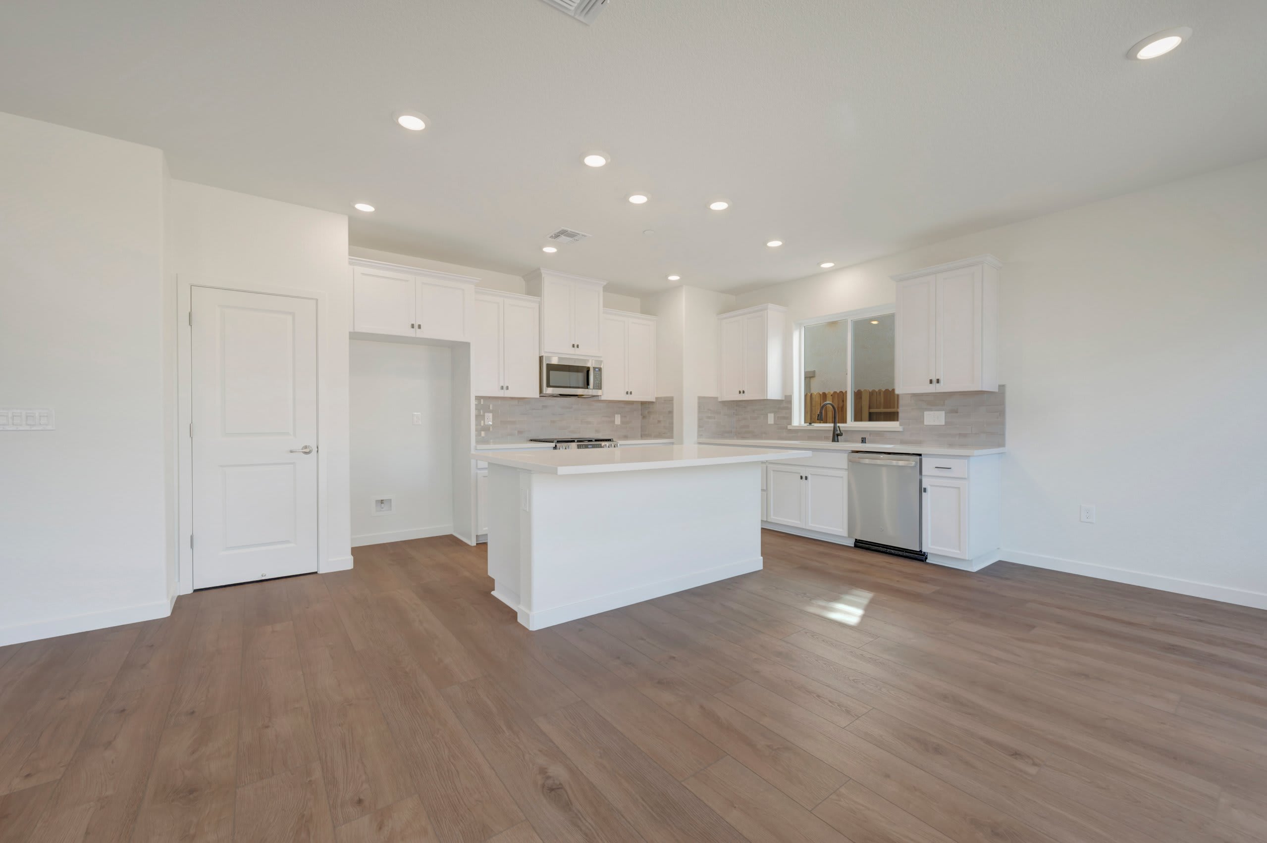 A modern, white kitchen with a large island and stainless steel appliances is visible in the background, while the foreground features a spacious room with light brown hardwood flooring.