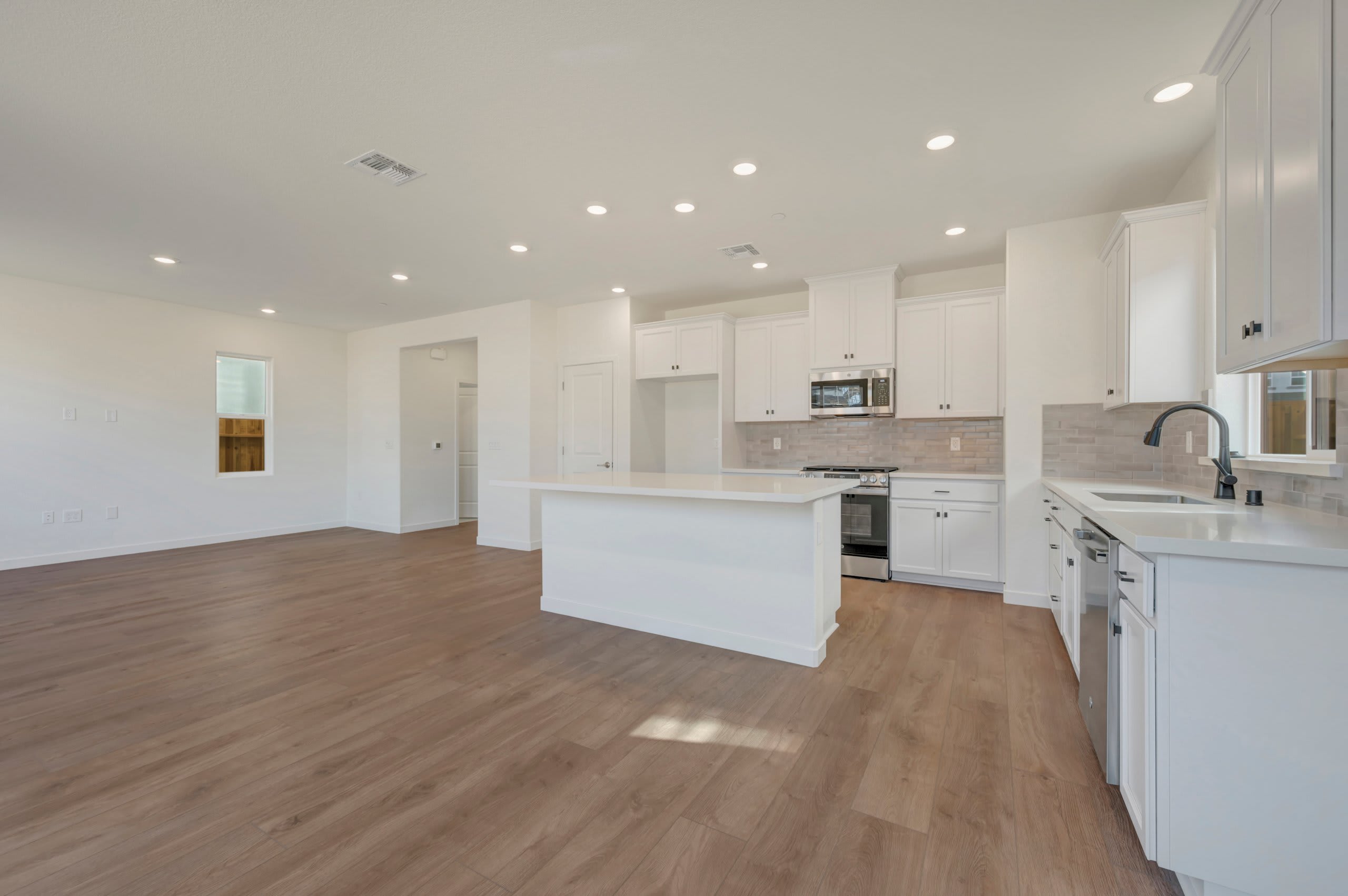 A modern, open-concept kitchen with white cabinets and a large island is in the foreground, while a spacious, empty living area with a window is in the background.