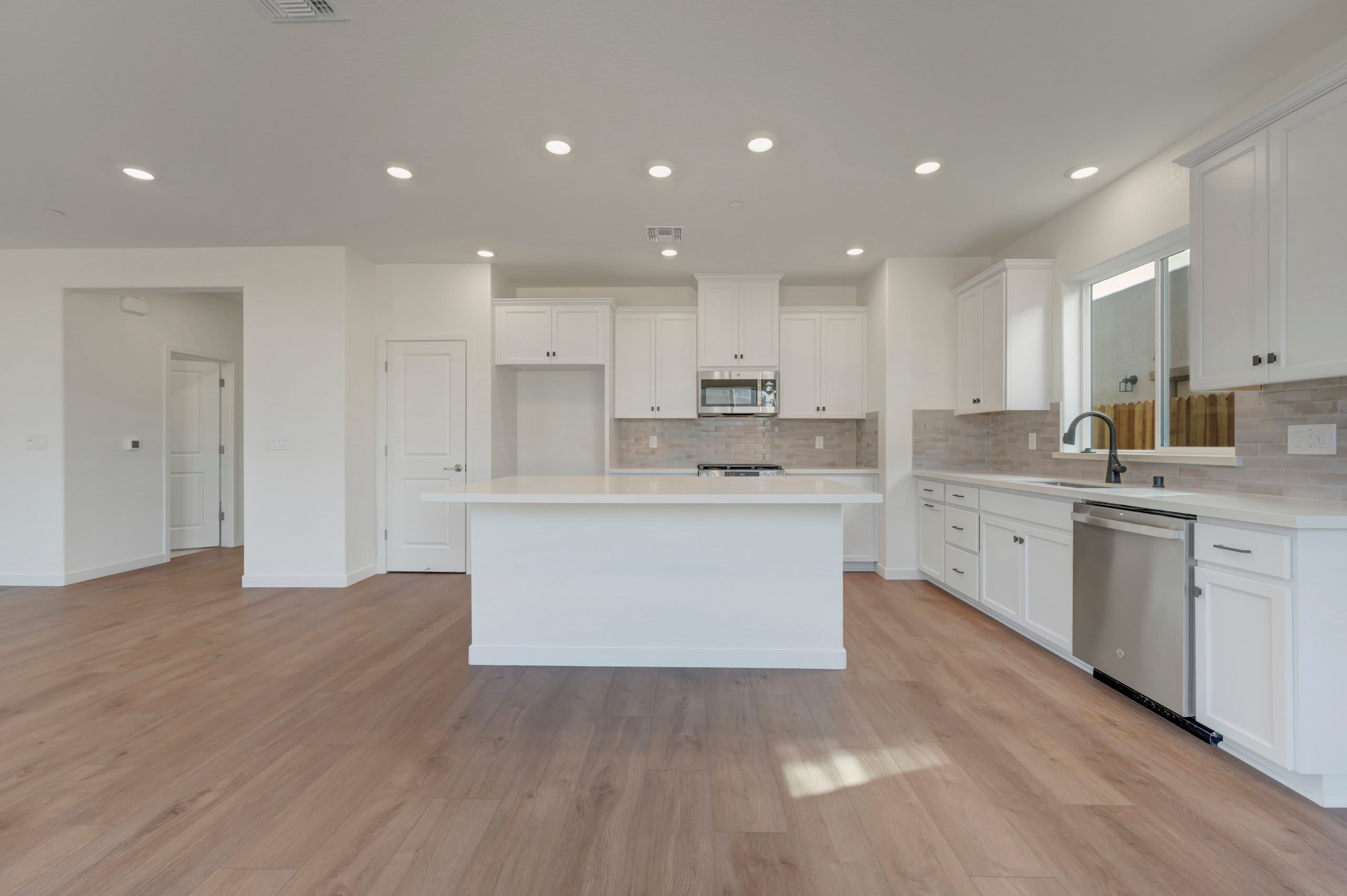 A modern white kitchen with a large island in the foreground and a hallway leading to another room in the background.