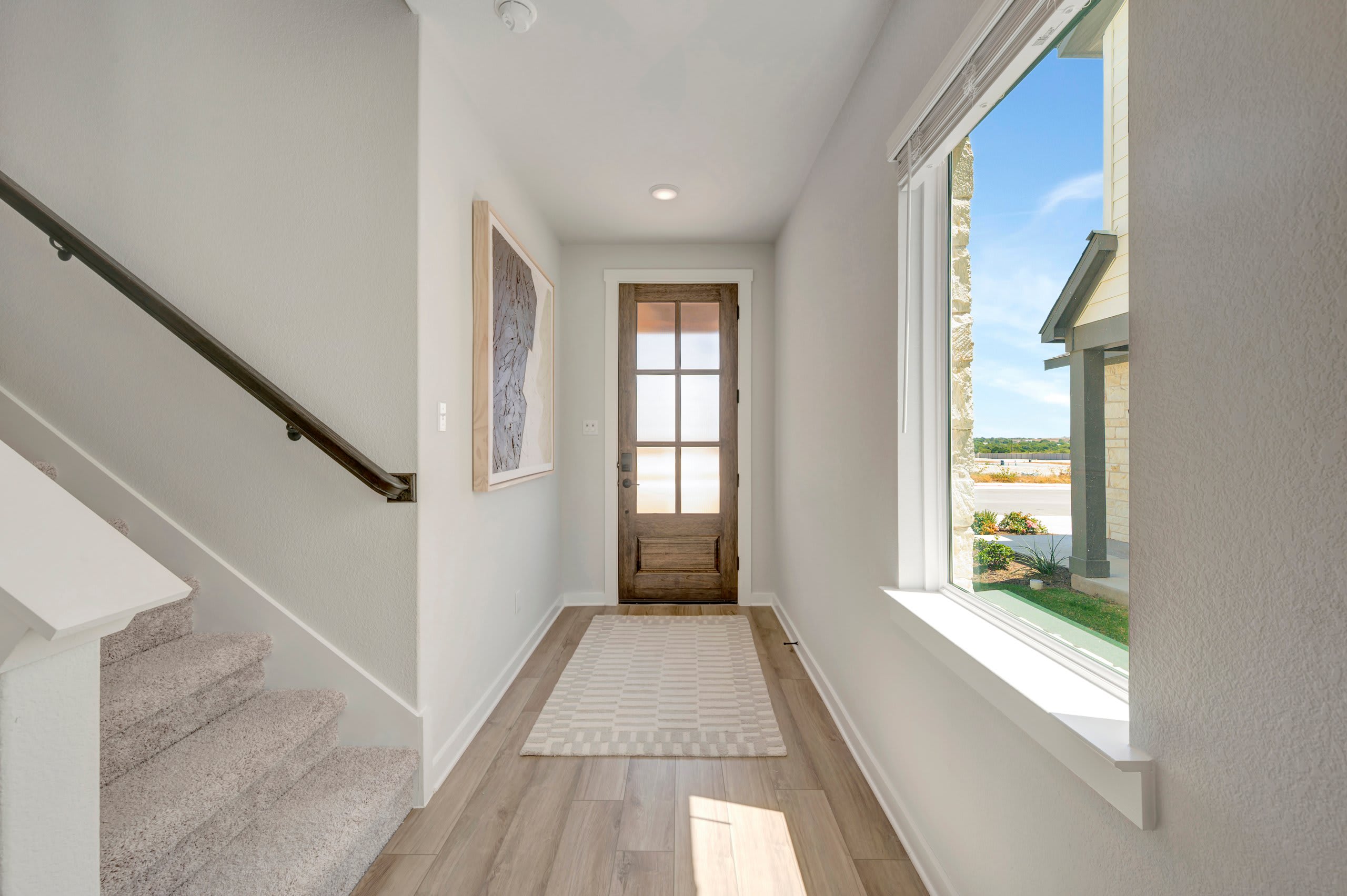 A bright entryway with a wooden front door and a striped rug in the foreground, and a large window revealing a sunny outdoor scene in the background.