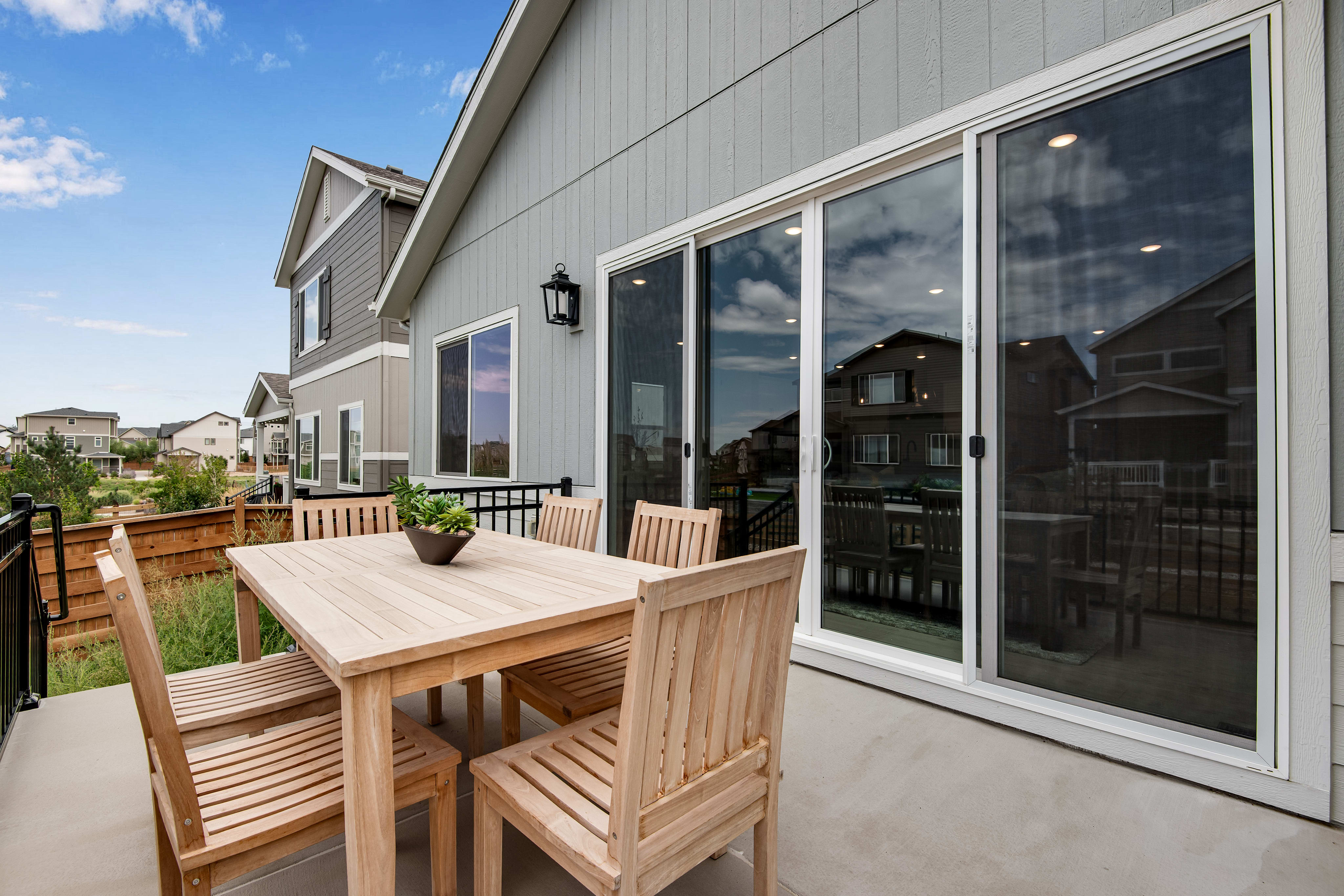 A wooden patio table and chairs sit on a concrete patio in front of a modern house with large sliding glass doors, while a suburban neighborhood stretches out in the background under a cloudy sky.