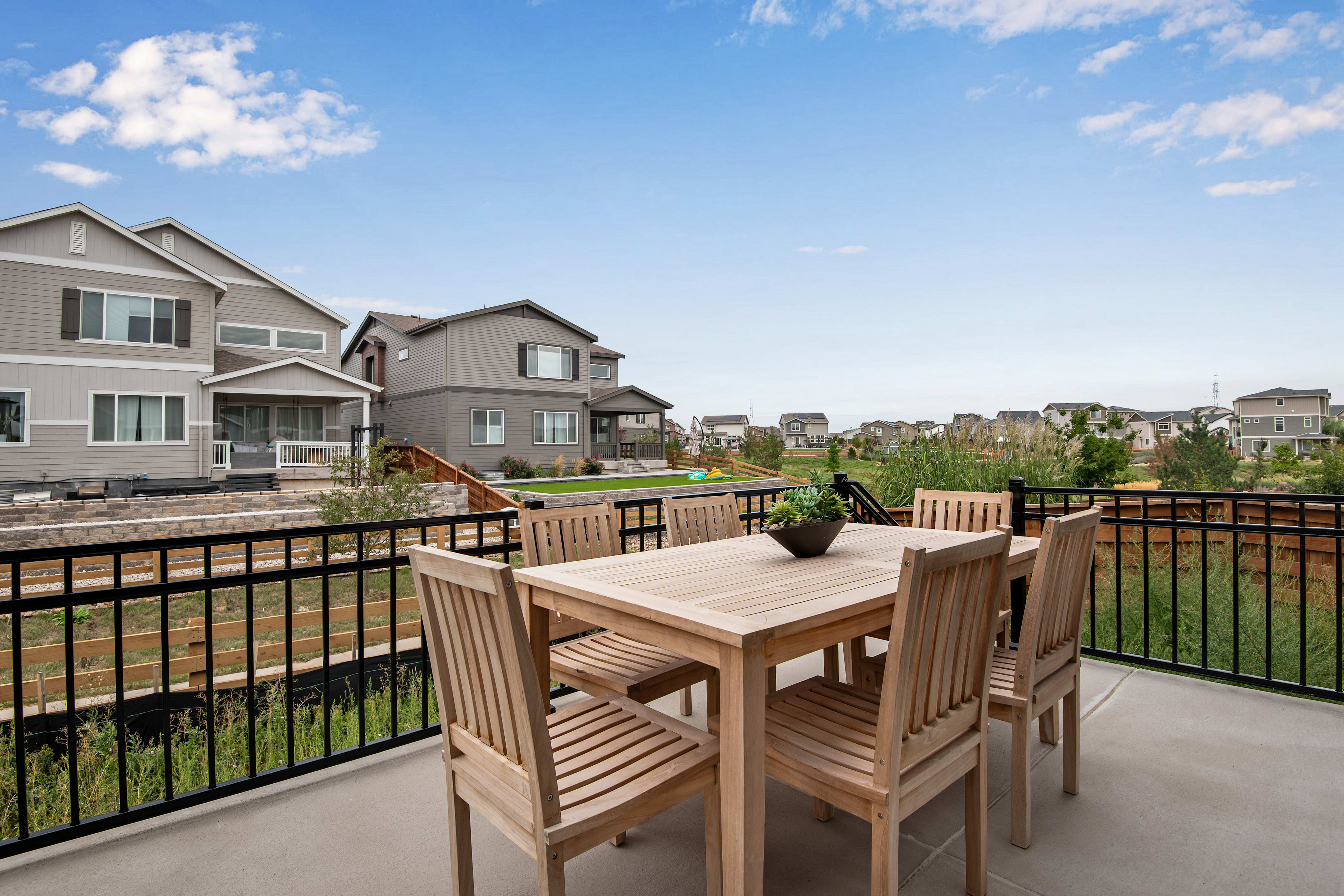 A wooden patio table and chairs sit on a concrete patio in the foreground, with a row of suburban houses and a bright blue sky with scattered clouds in the background.