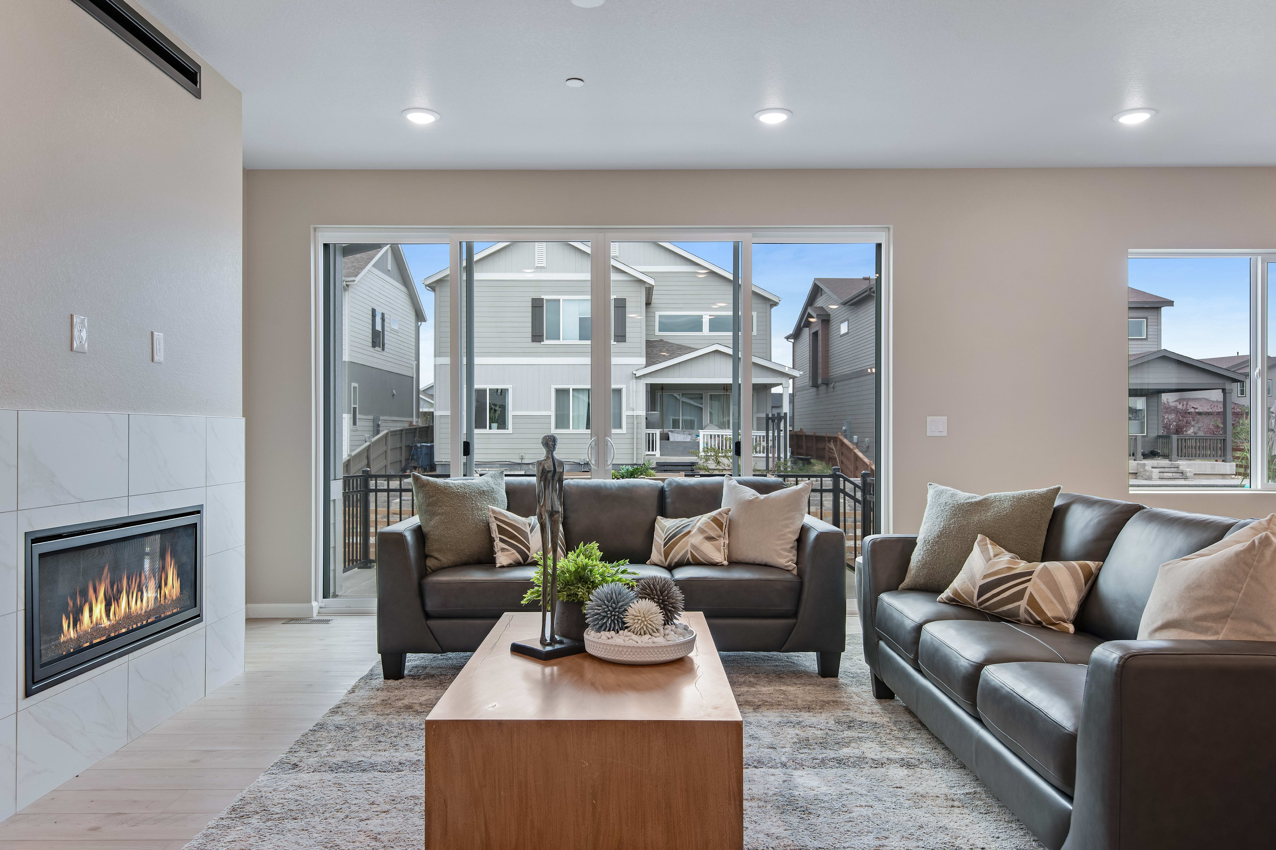 A modern living room with a fireplace and leather sofas in the foreground looks out onto a suburban neighborhood through large glass doors in the background.