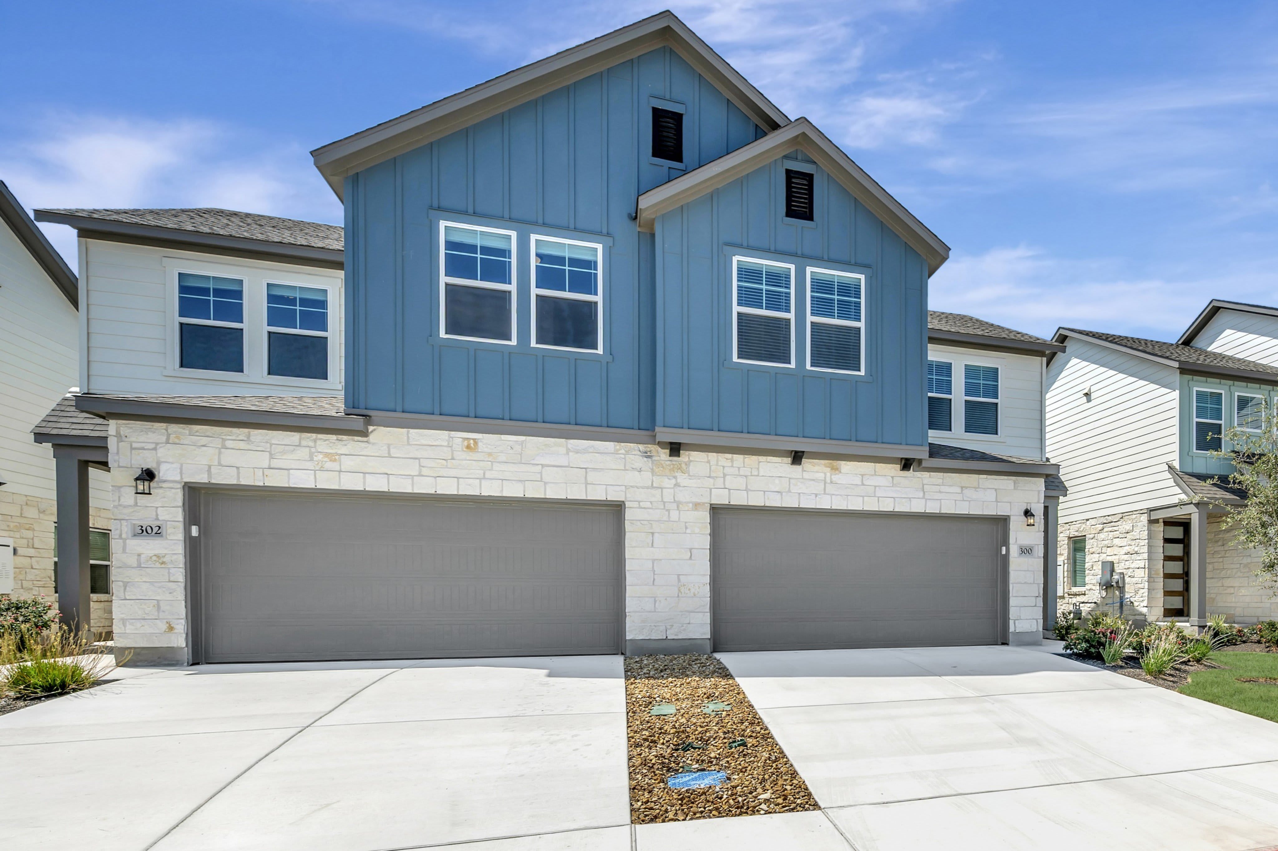 A modern blue and white townhouse with two garage doors in the foreground sits under a clear blue sky with fluffy clouds.