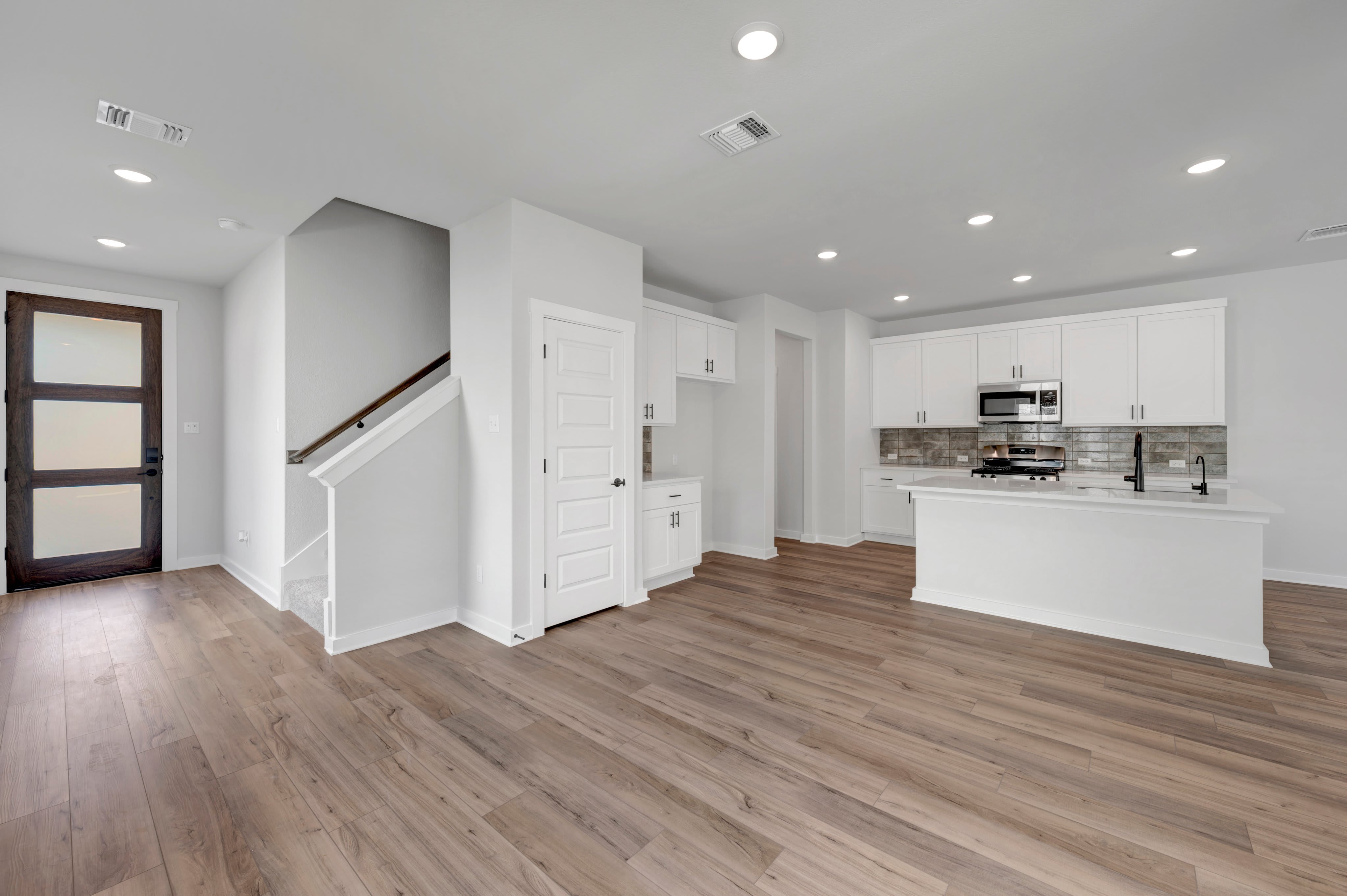 A modern open-concept living space features a dark wood front door and staircase in the foreground, leading to a sleek white kitchen with an island in the background.