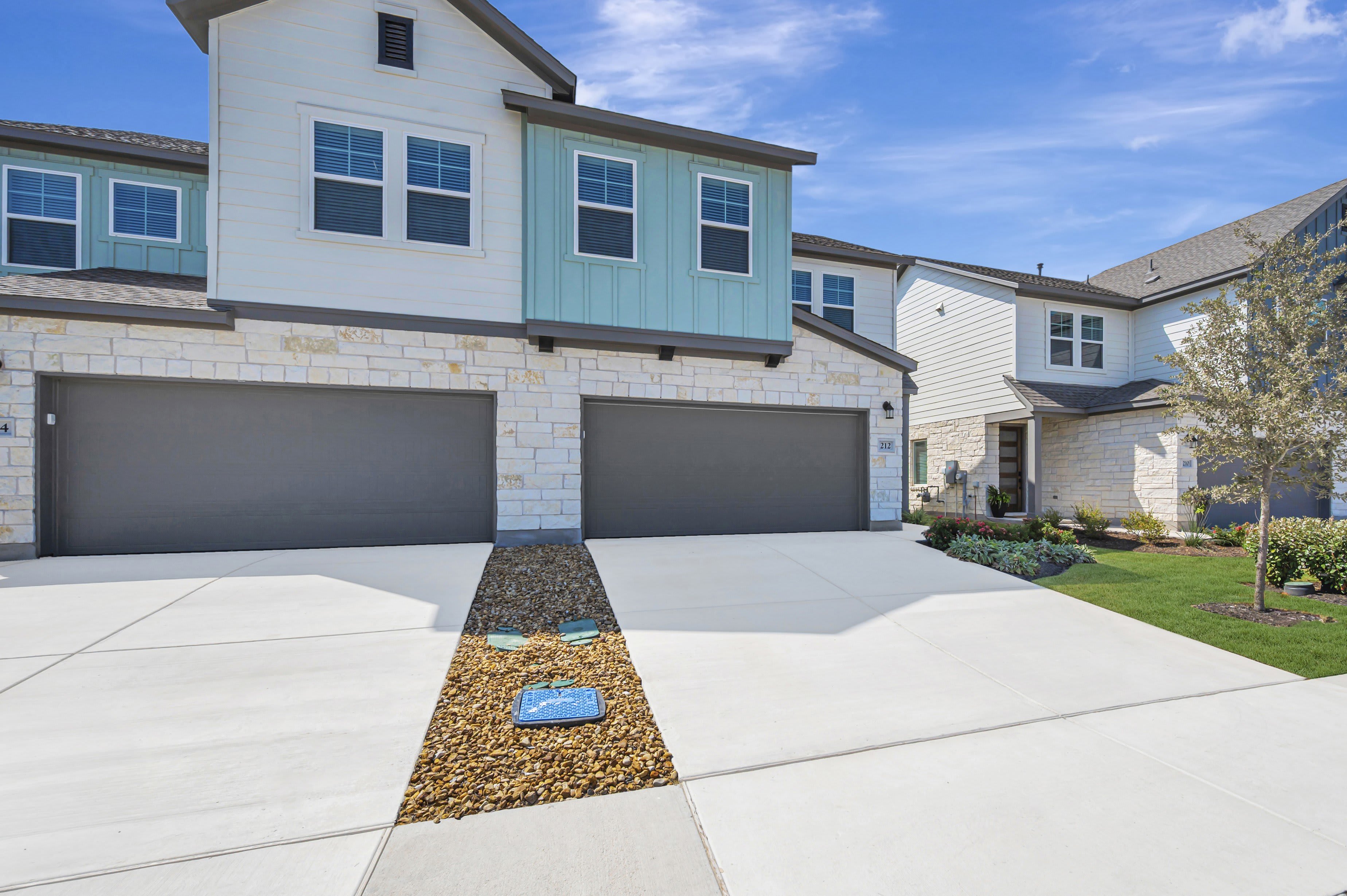 A modern townhouse complex with light-colored stone and blue siding is visible in the background, while a concrete driveway with a small rock garden is in the foreground.