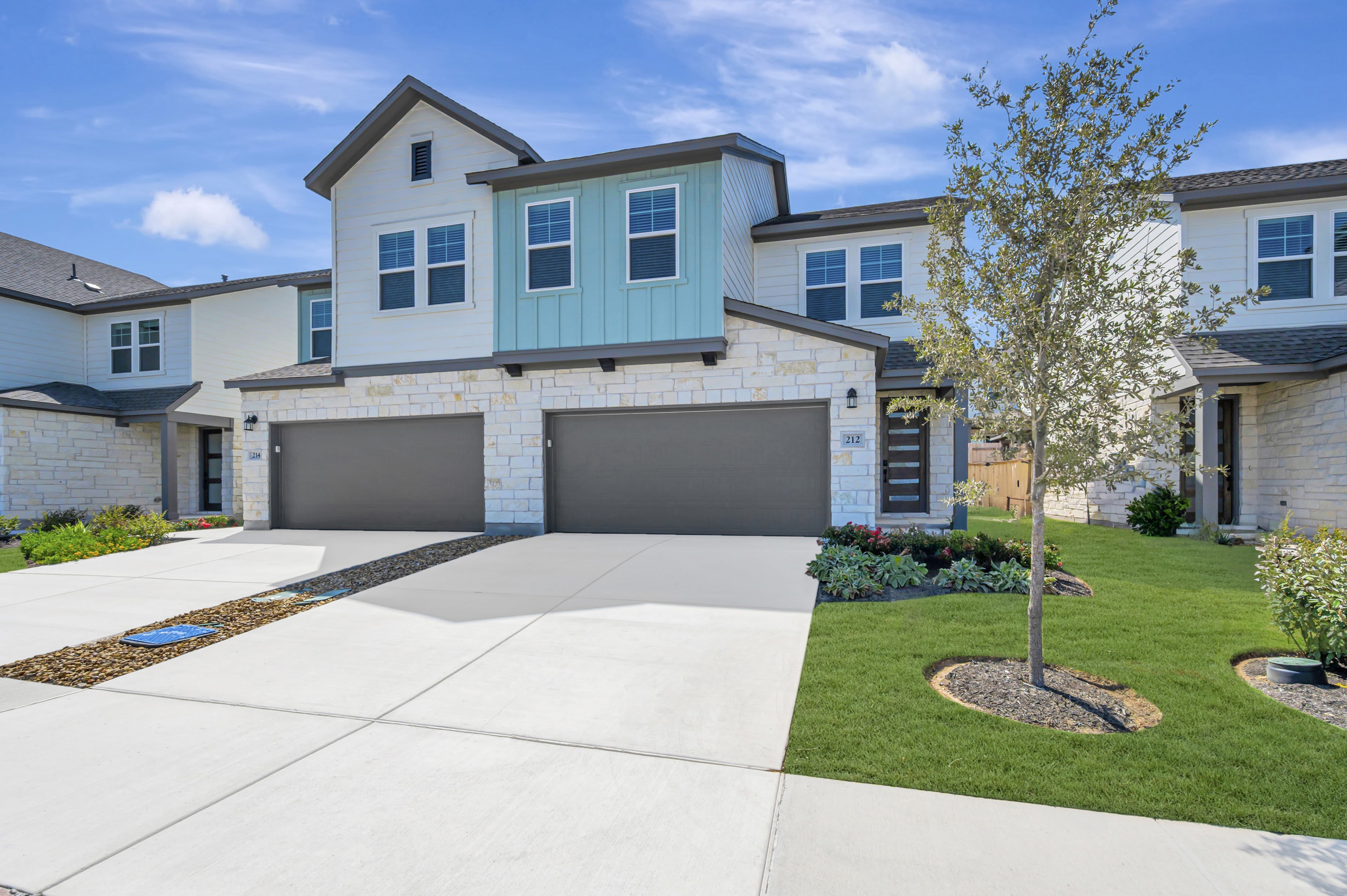 A modern townhouse with a light-colored stone facade and blue accents stands on a manicured lawn with a paved driveway in the foreground, under a bright blue sky with scattered clouds.