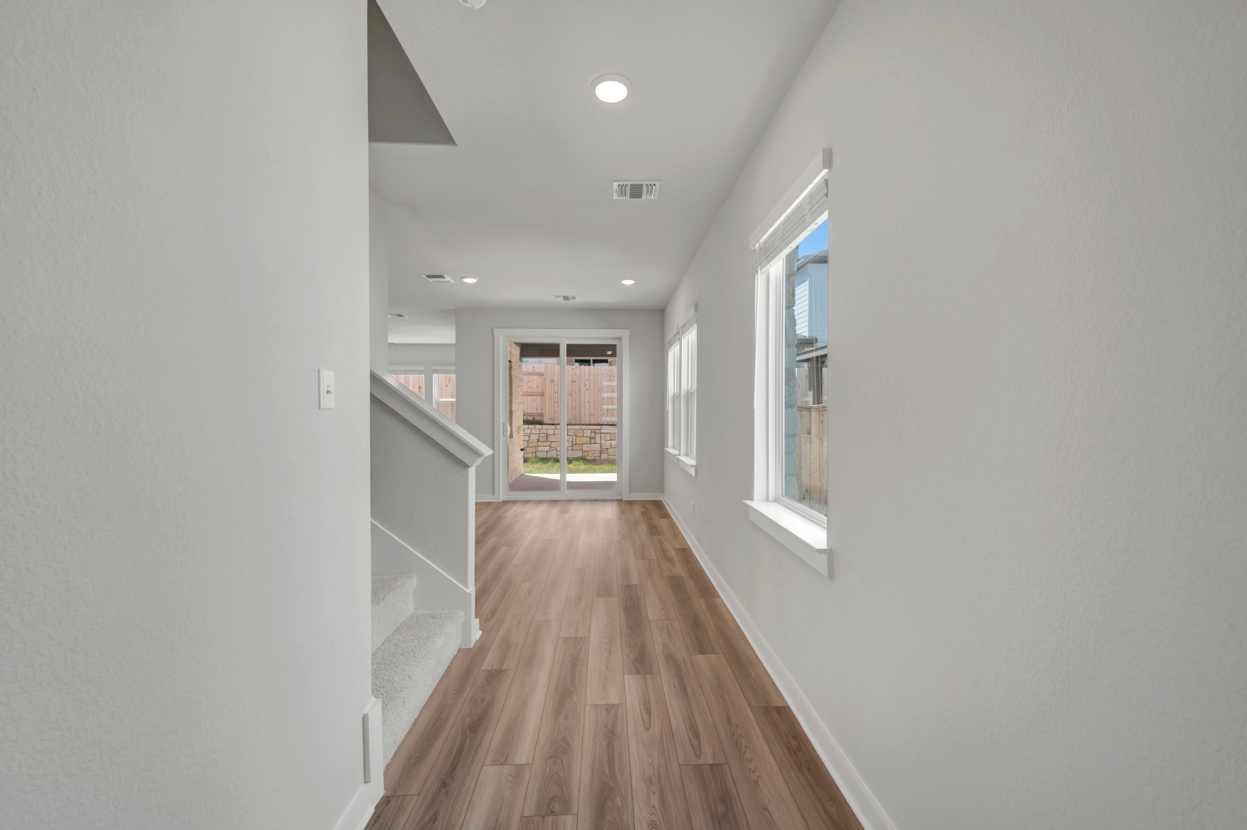 A bright hallway with a staircase on the left leads to a living area with large windows and a sliding glass door opening to a backyard.