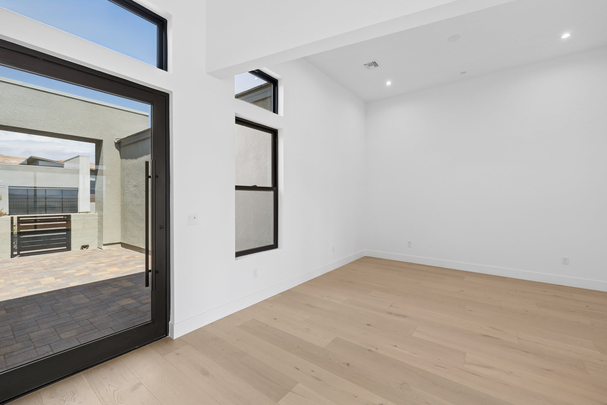 A modern, empty room with light wood floors and white walls opens to a patio with dark paving stones through a large glass door.