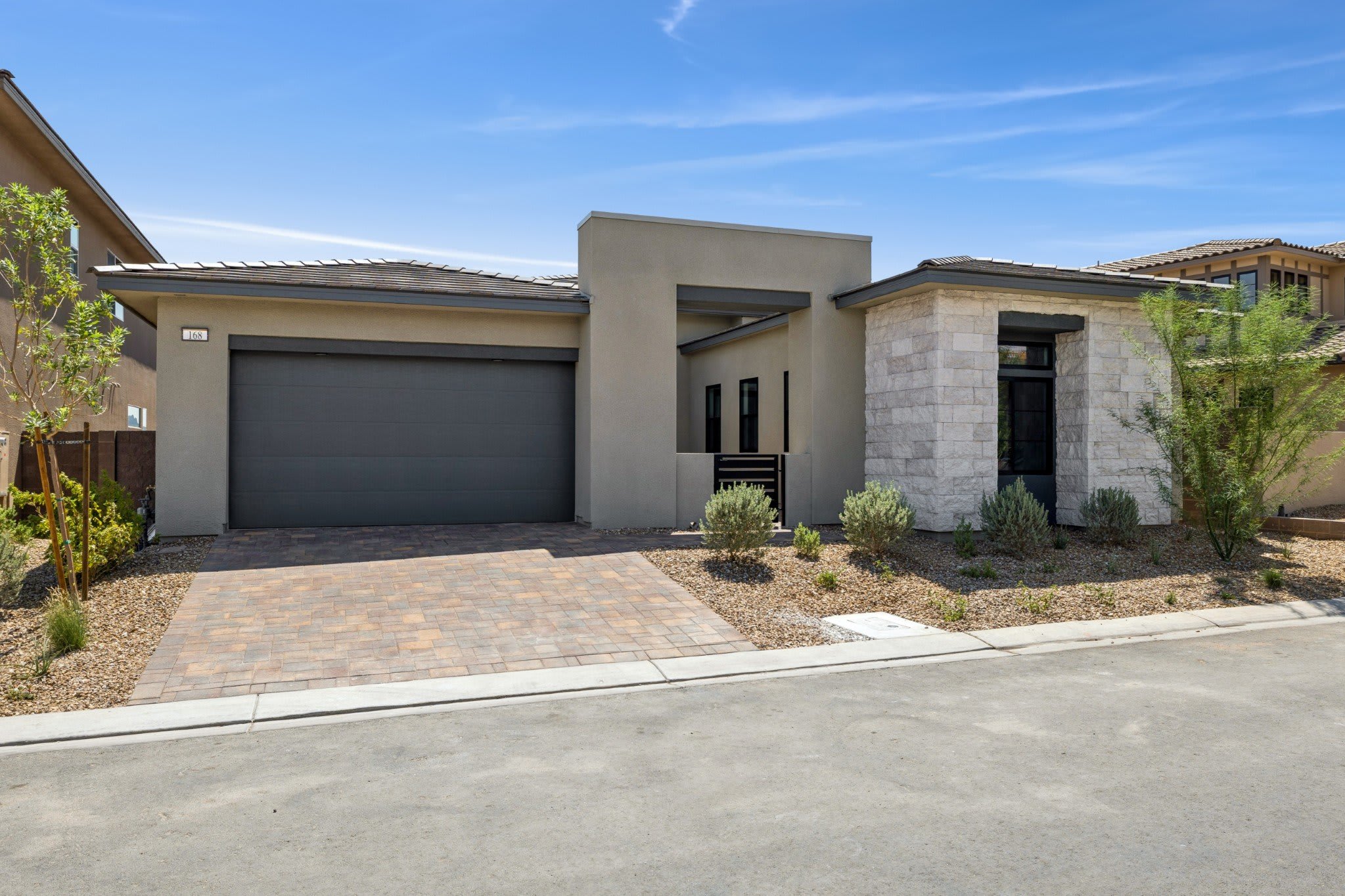 A modern beige and stone house with a paved driveway sits under a bright blue sky with scattered clouds.