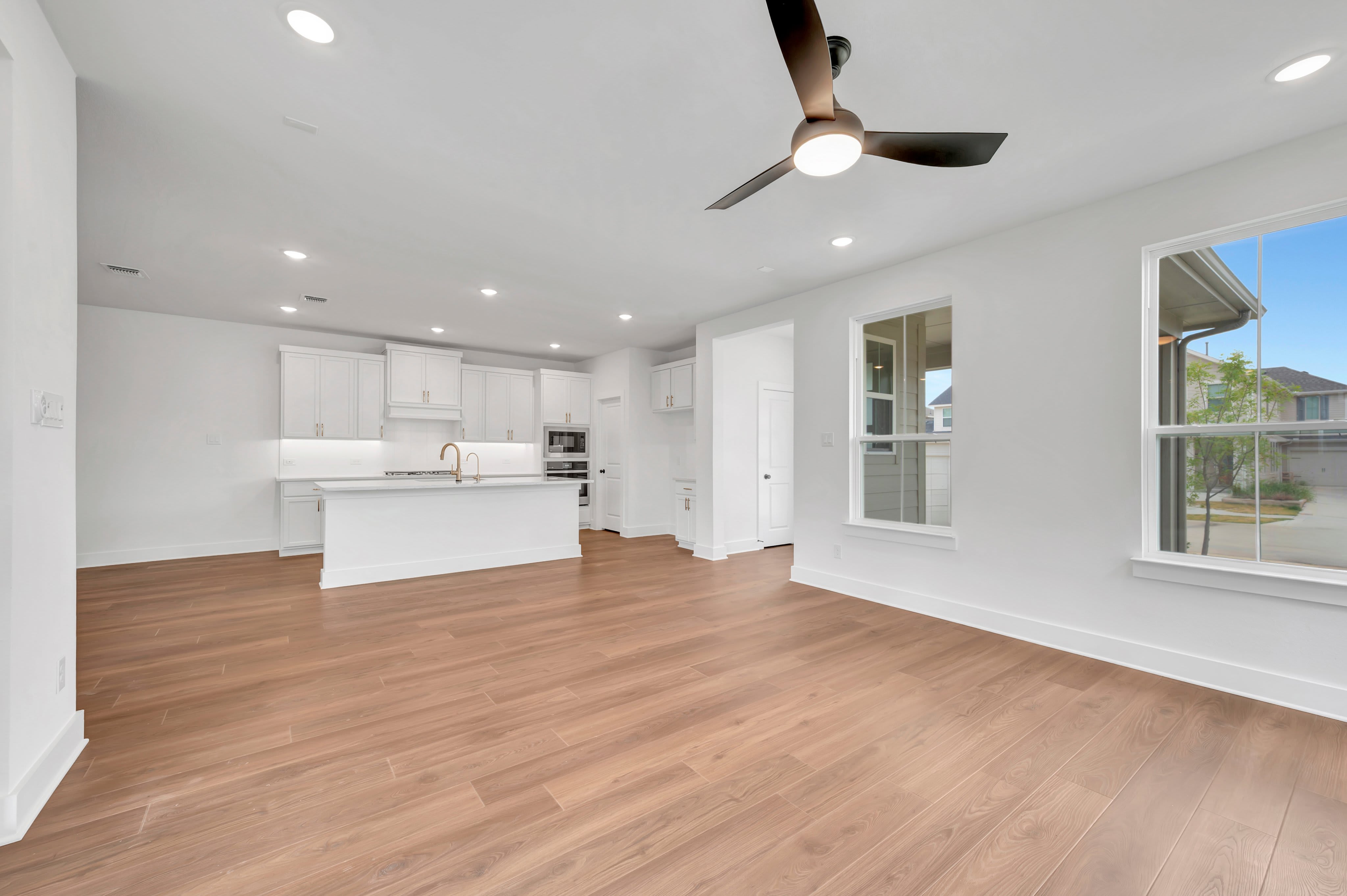 A spacious, modern living area with a light wood floor in the foreground opens to a white kitchen with an island in the background, while large windows offer views of a suburban exterior.