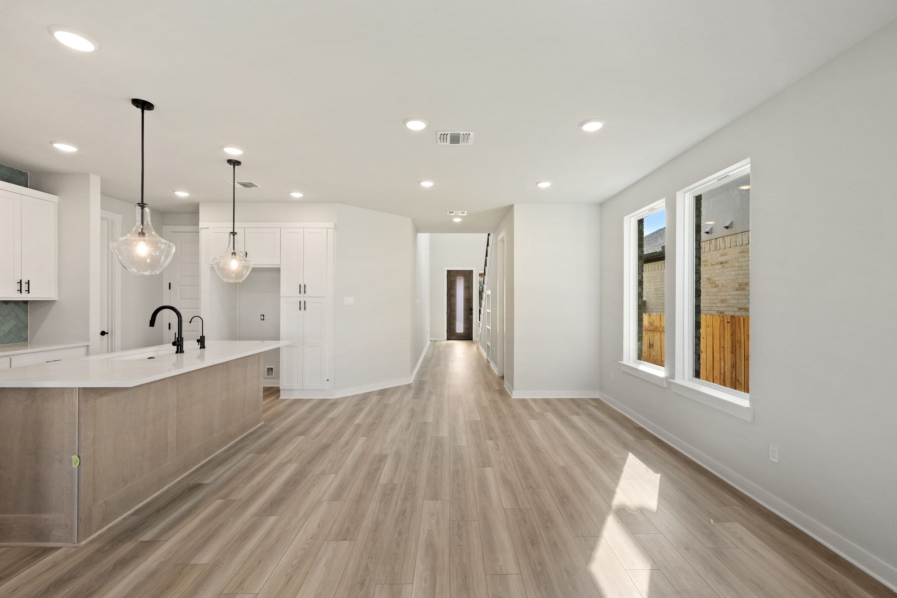 A modern kitchen island with pendant lights is in the foreground, while a hallway leading to a staircase and windows overlooking a backyard are in the background.