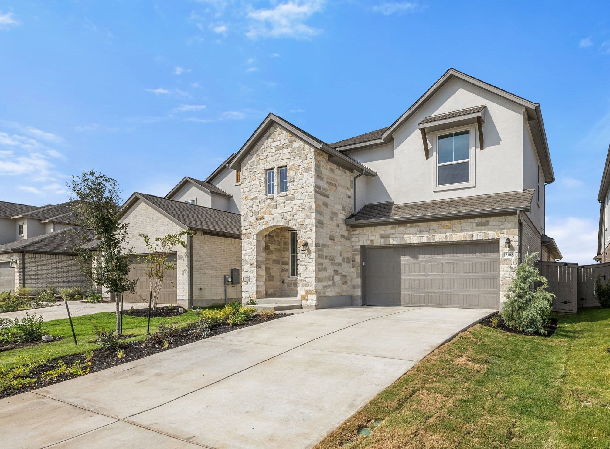 A modern stone and stucco house with a concrete driveway in the foreground sits under a clear blue sky with other houses visible in the background.