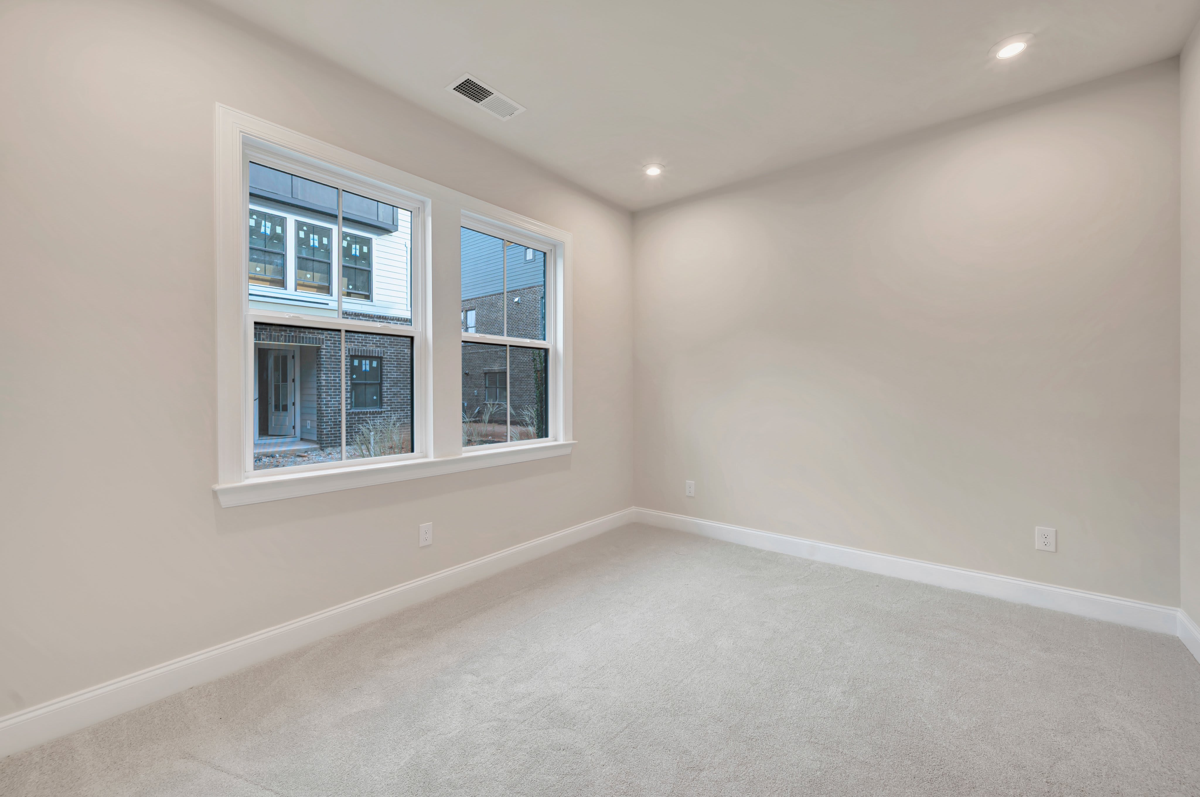 A carpeted room with two windows looks out onto a brick building.