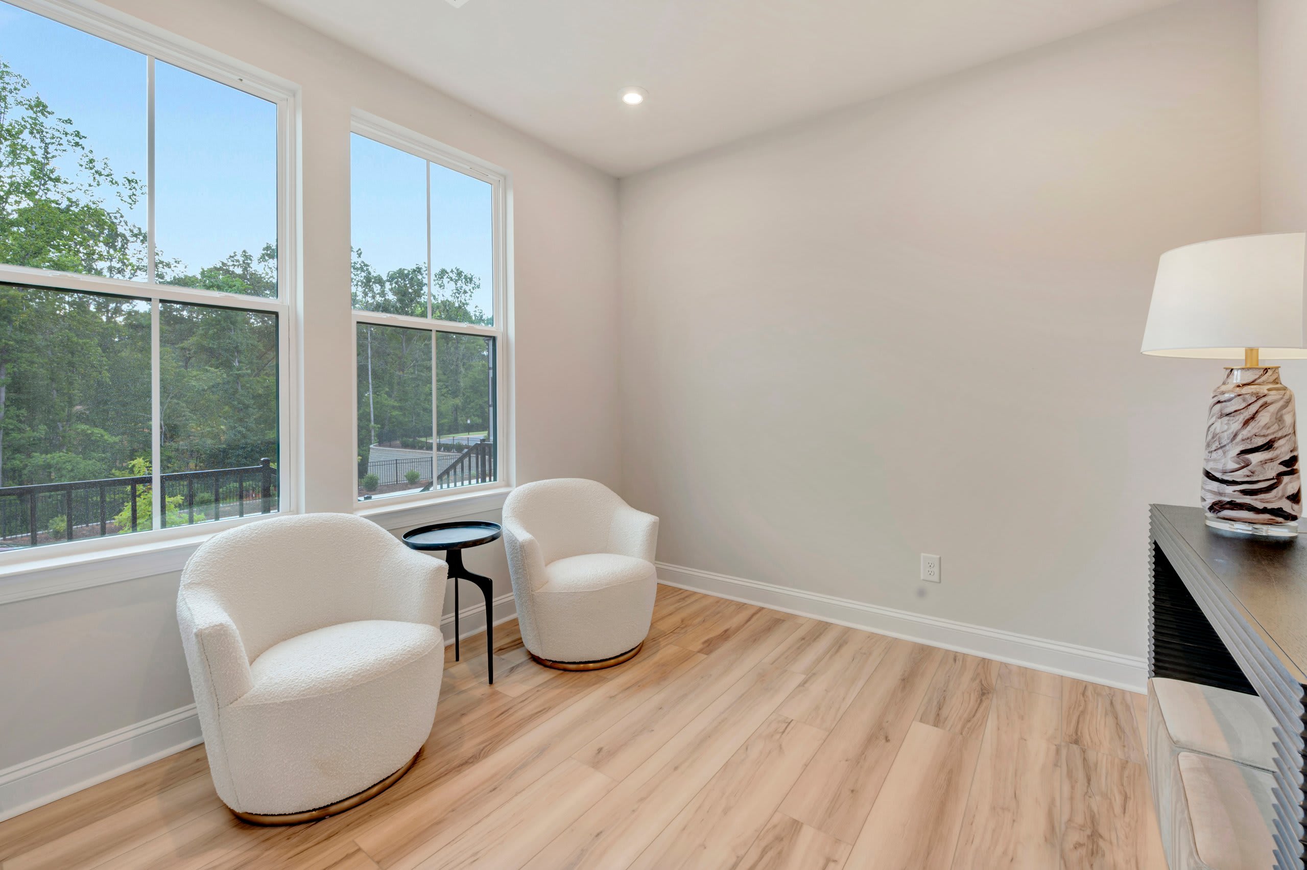 Two white armchairs and a small black table sit on a light wood floor in the foreground, while large windows reveal a lush green forest in the background.