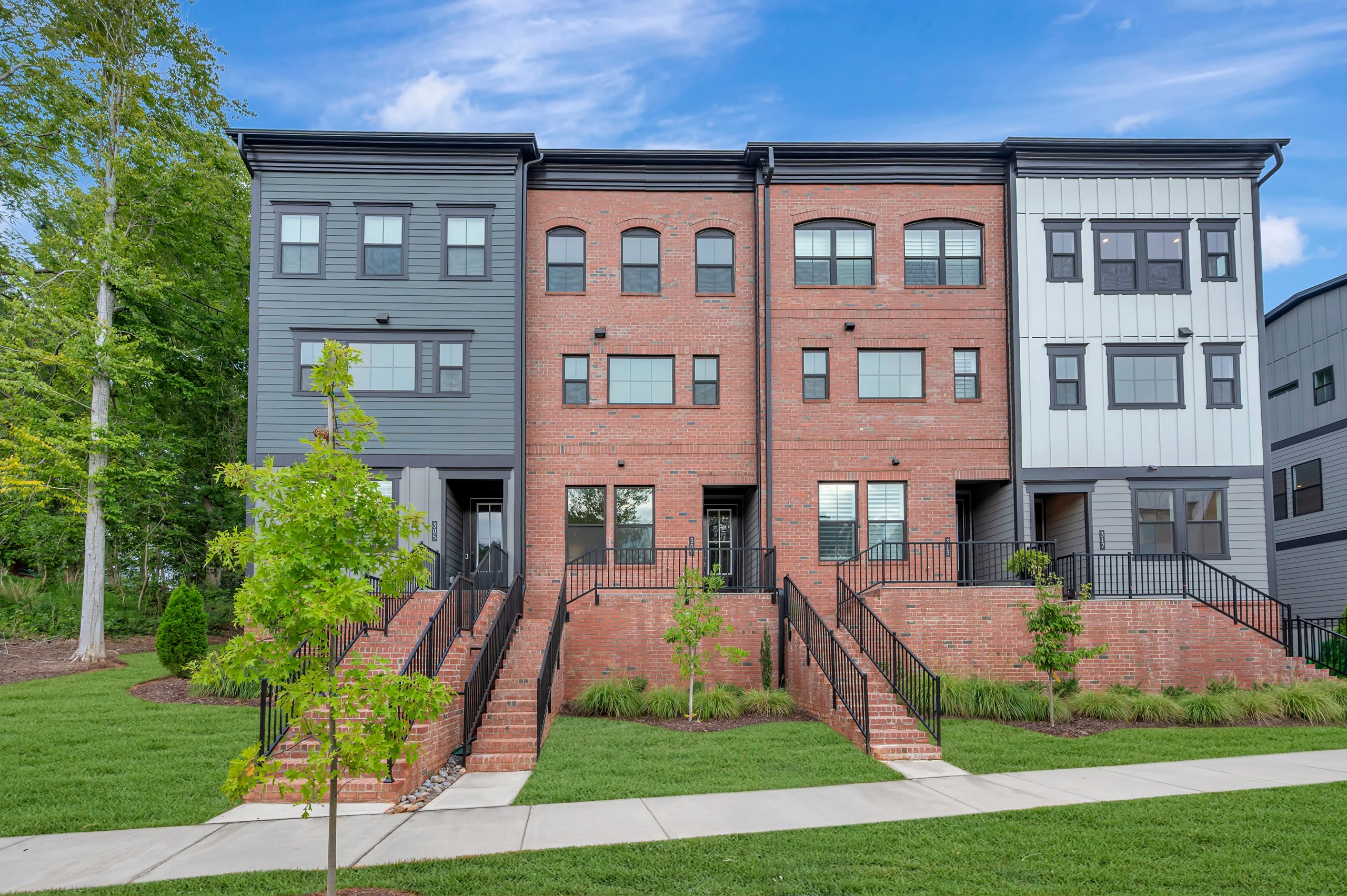 A row of modern townhouses with brick and siding exteriors stands against a backdrop of lush green trees and a bright blue sky.