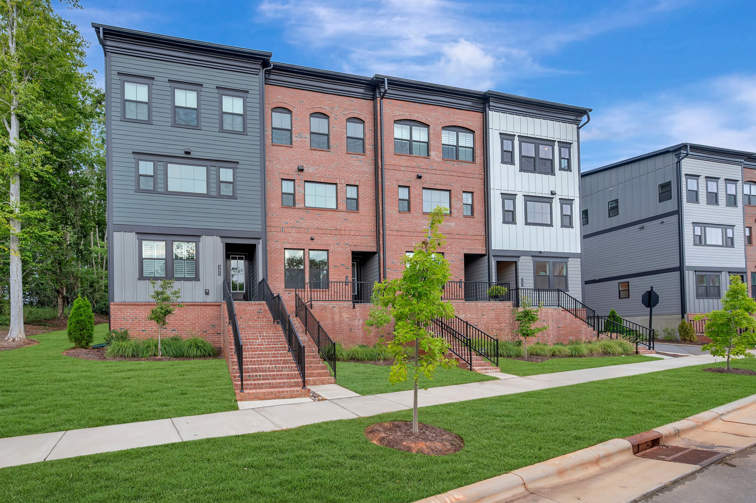 A row of modern townhouses with brick and gray siding stands on a manicured lawn under a blue sky with scattered clouds.