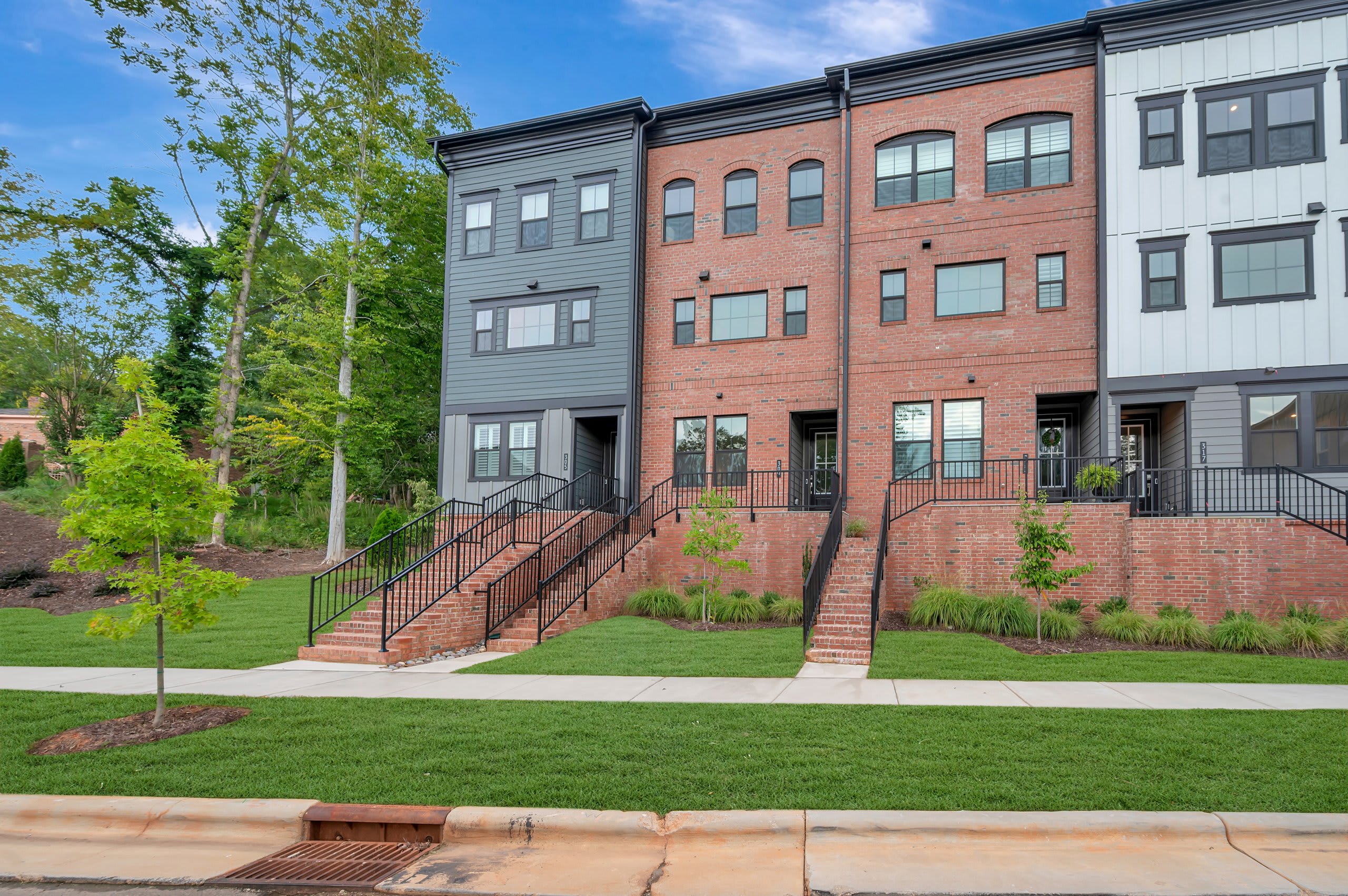 A manicured lawn and sidewalk lead to a row of modern townhouses with brick and gray exteriors, backed by lush green trees under a cloudy sky.
