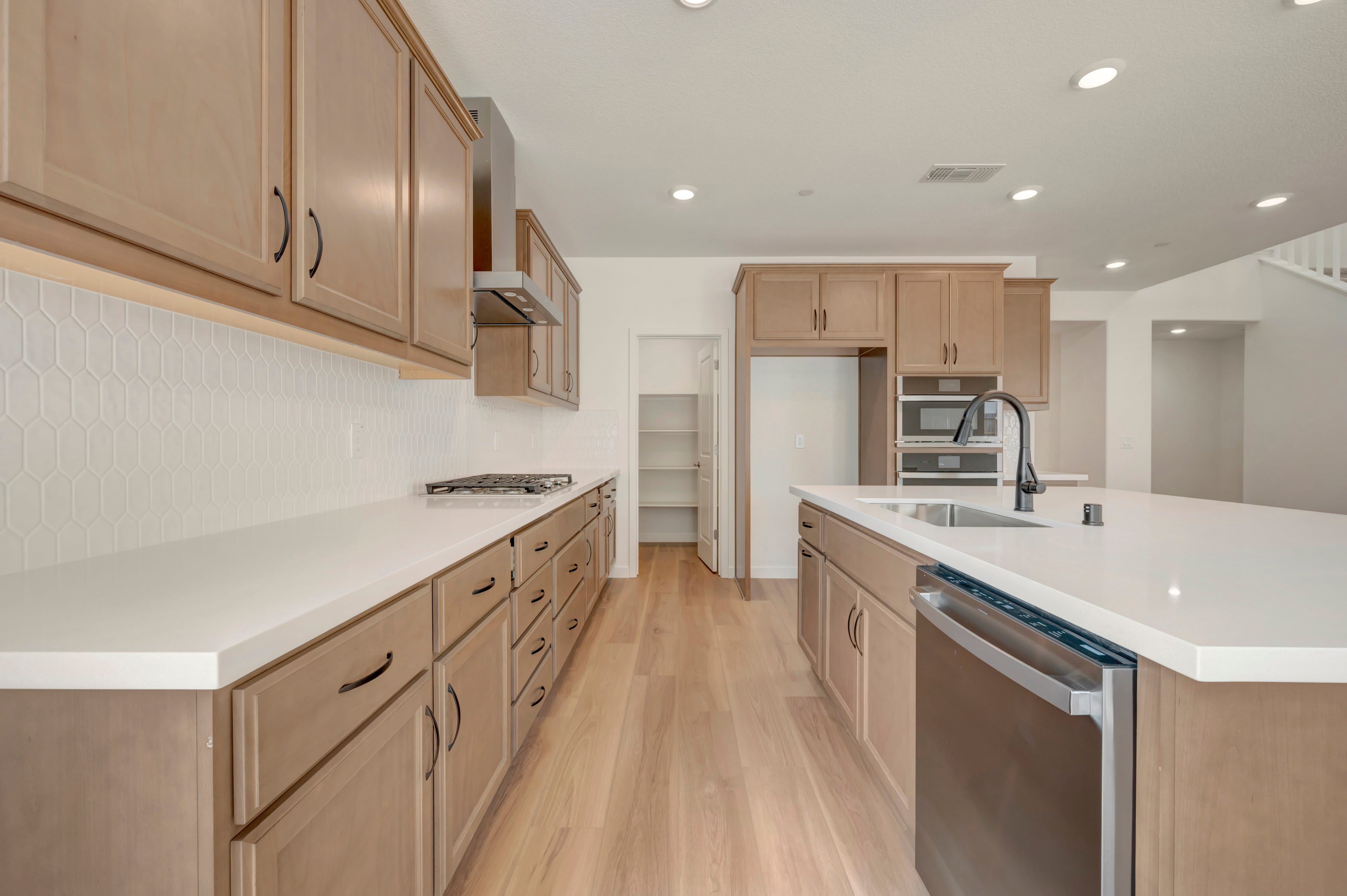 A modern kitchen with light wood cabinets and white countertops features a large island in the foreground and a pantry and staircase in the background.