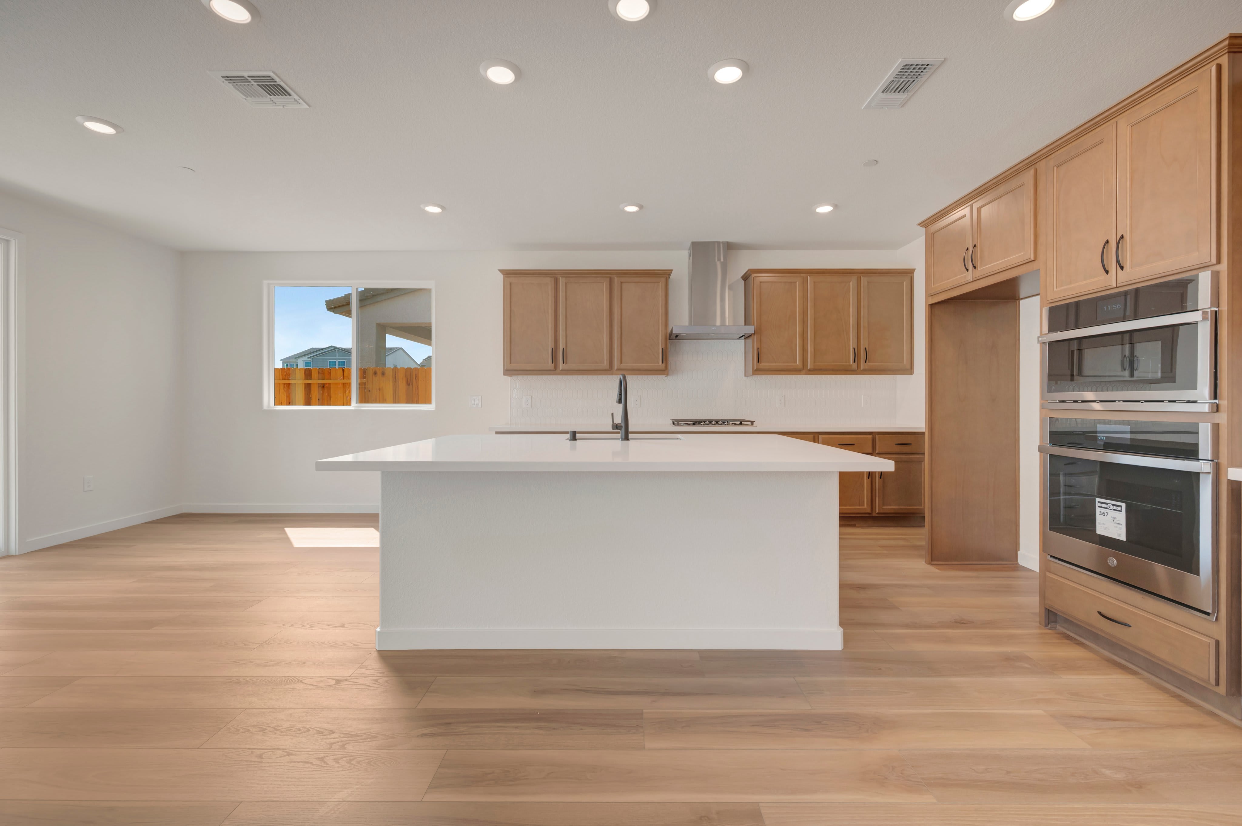 A modern kitchen with a white island in the foreground and light wood cabinets and stainless steel appliances in the background, with a window showing a glimpse of the outdoors.