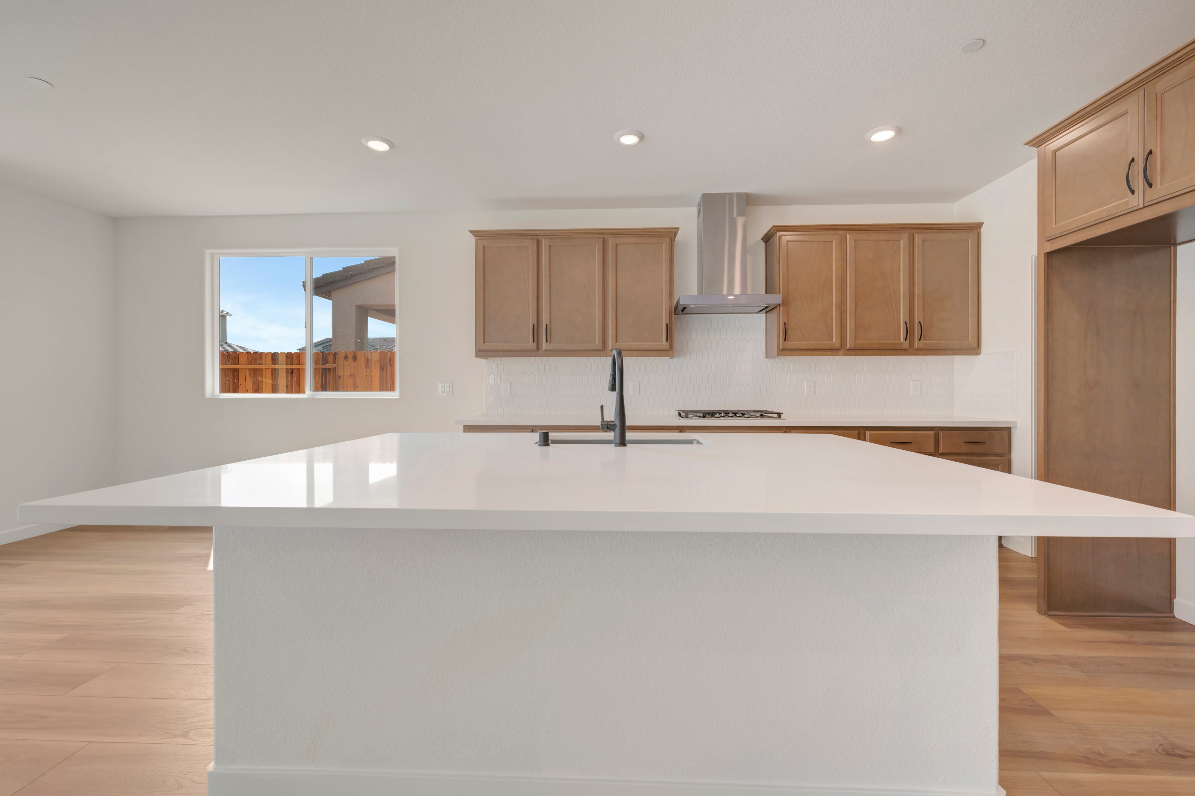 A modern kitchen features a large white island in the foreground and light wood cabinets and stainless steel appliances in the background, with a window showing a fence and blue sky.