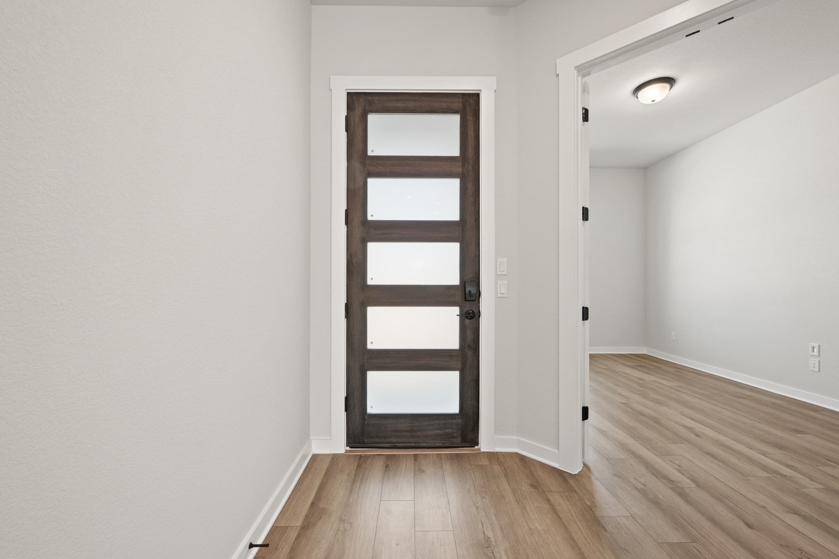 A dark wood door with five frosted glass panels stands in the foreground, while a brightly lit room with wood flooring is visible through an open doorway in the background.