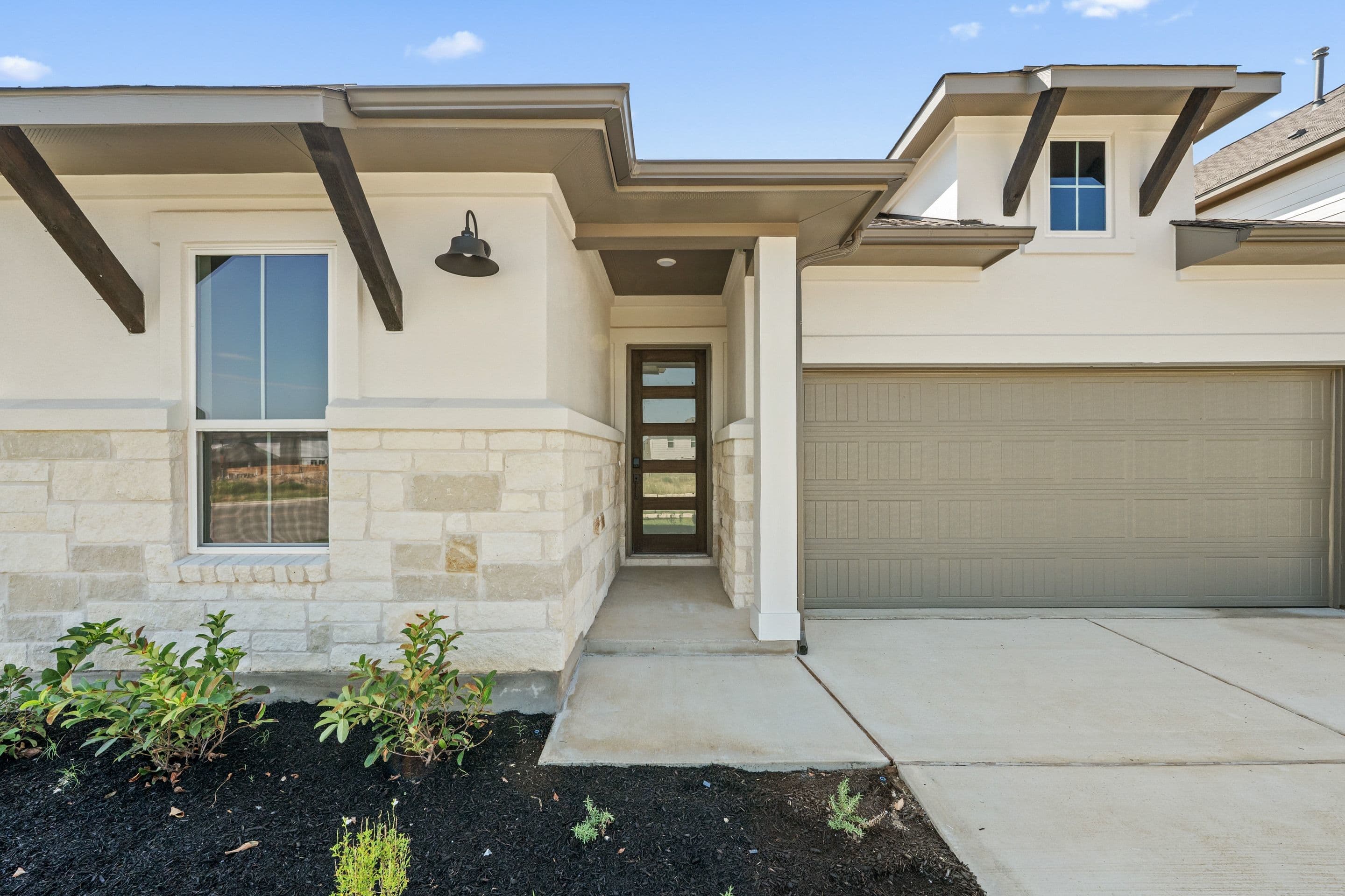 A modern house with a stone and stucco facade, a glass-paneled front door, and a beige garage door is set against a clear blue sky.