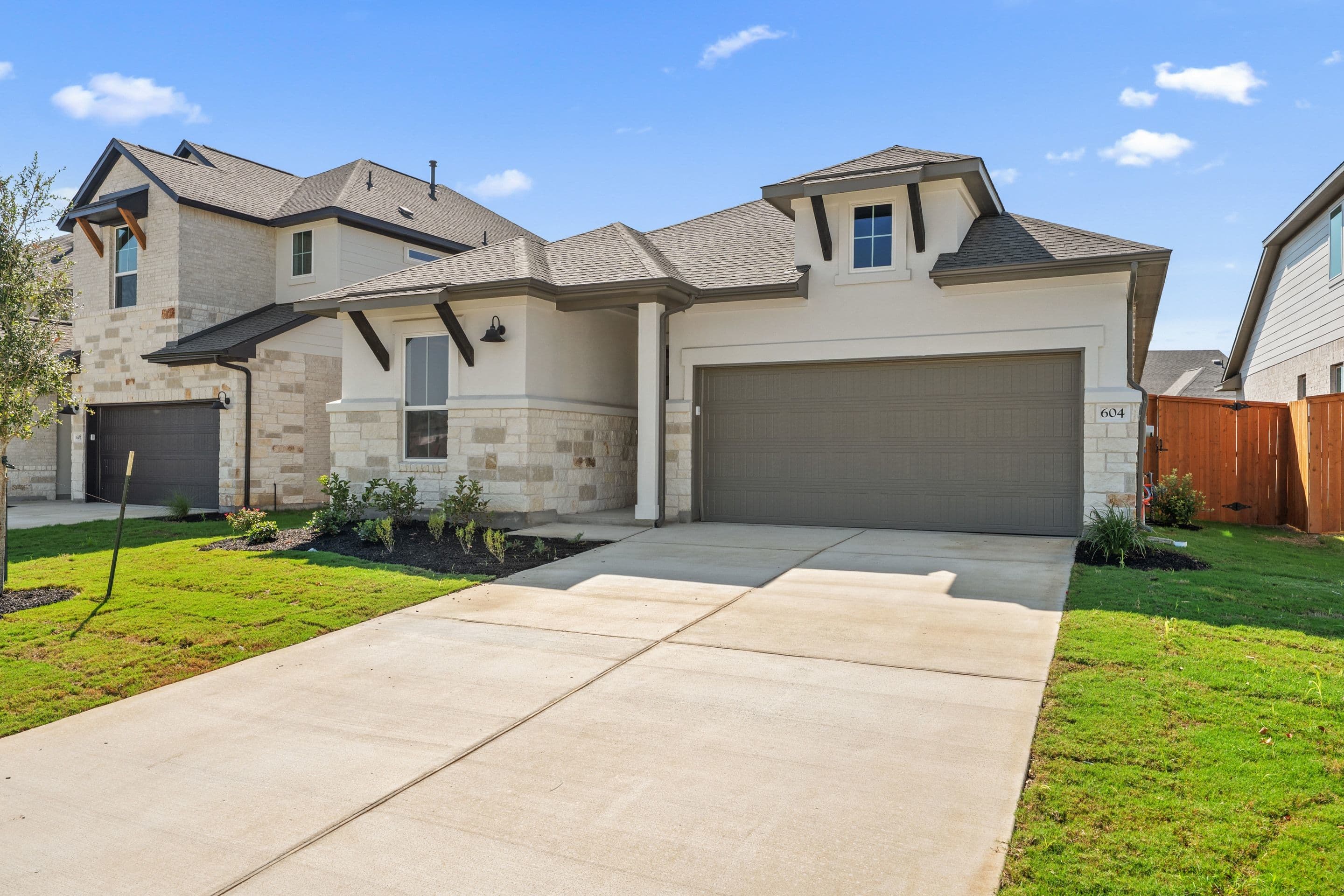 A modern stucco and stone house with a large garage door sits on a manicured lawn, with other similar houses visible in the background under a clear blue sky.