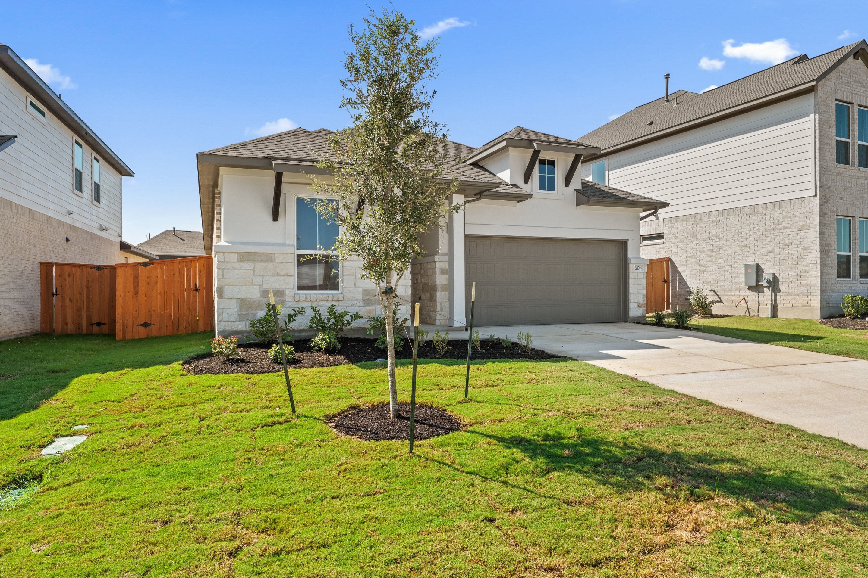 A modern house with a manicured lawn in the foreground sits under a bright blue sky with fluffy clouds, while neighboring houses are visible in the background.