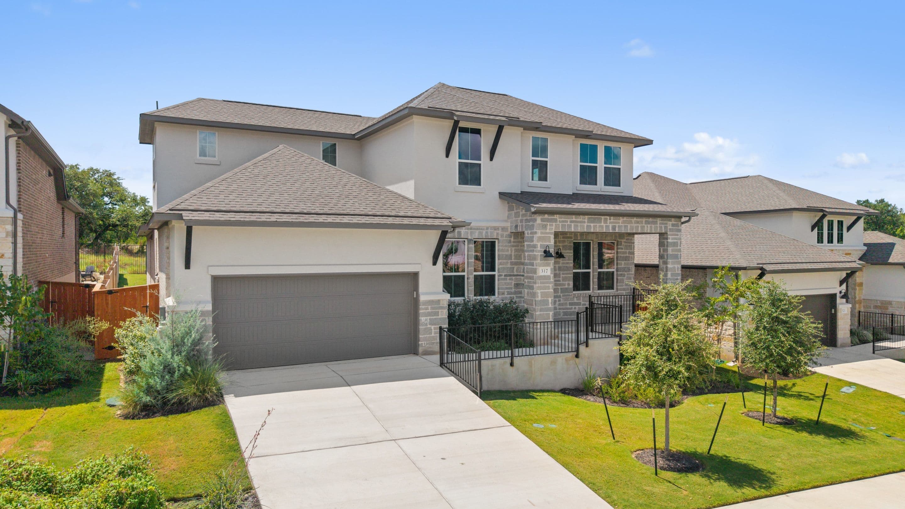 A modern two-story house with a gray garage door and stone accents sits on a manicured lawn, with neighboring houses and a clear blue sky in the background.