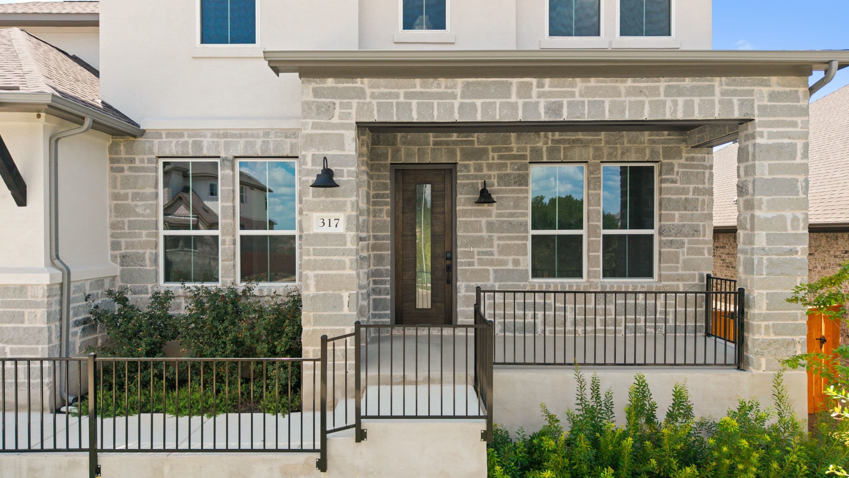 A modern stone-clad house with a glass-paneled front door and black metal railings in the foreground is set against a clear blue sky.