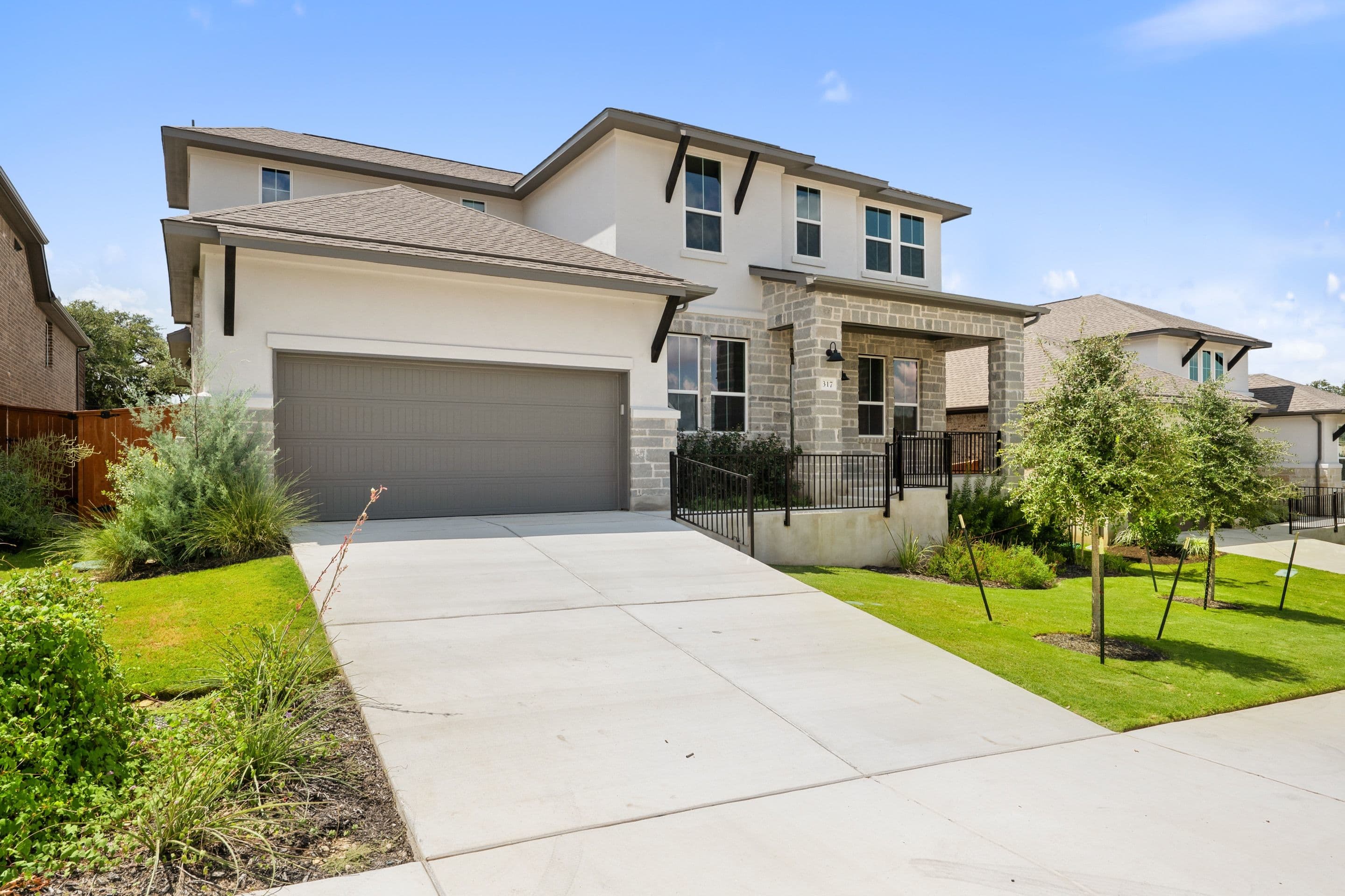 A modern two-story house with a gray garage door and stone accents sits on a concrete driveway, with a clear blue sky and neighboring houses in the background.