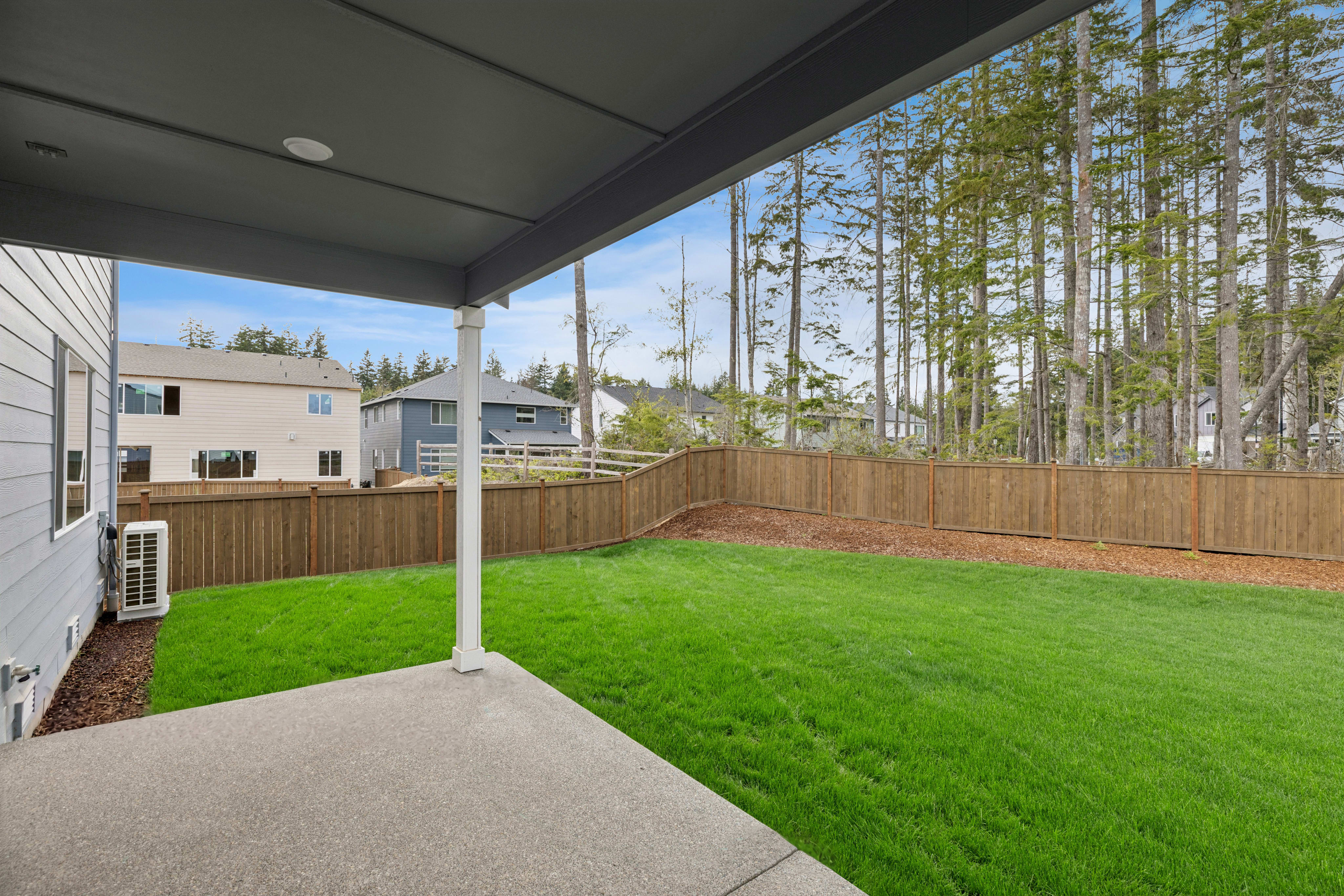 A covered patio overlooks a lush green lawn and a wooden fence, with houses and a dense forest in the background.