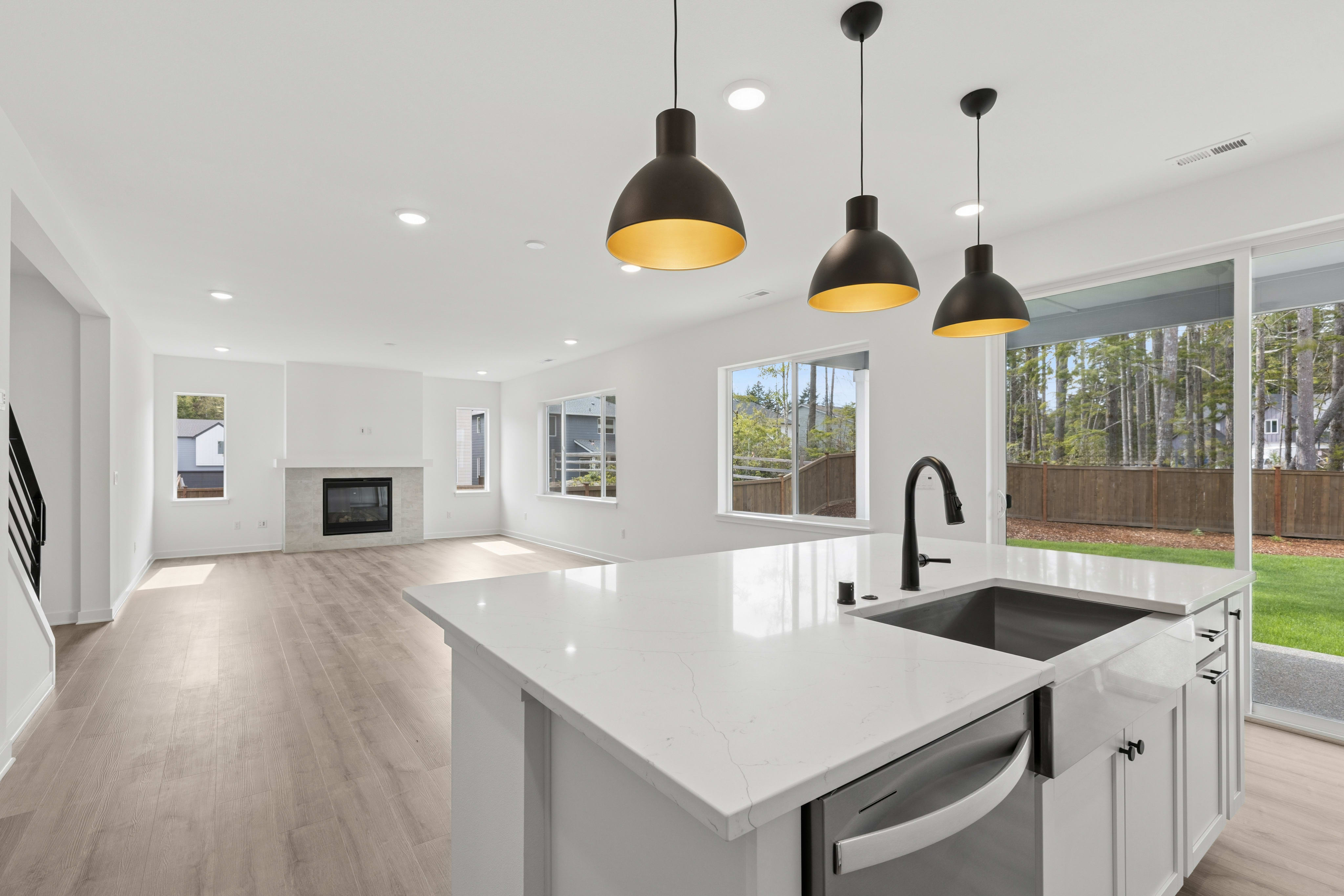 A modern kitchen island with a sink and pendant lights is in the foreground, while a spacious, brightly lit living area with a fireplace and large windows is in the background.