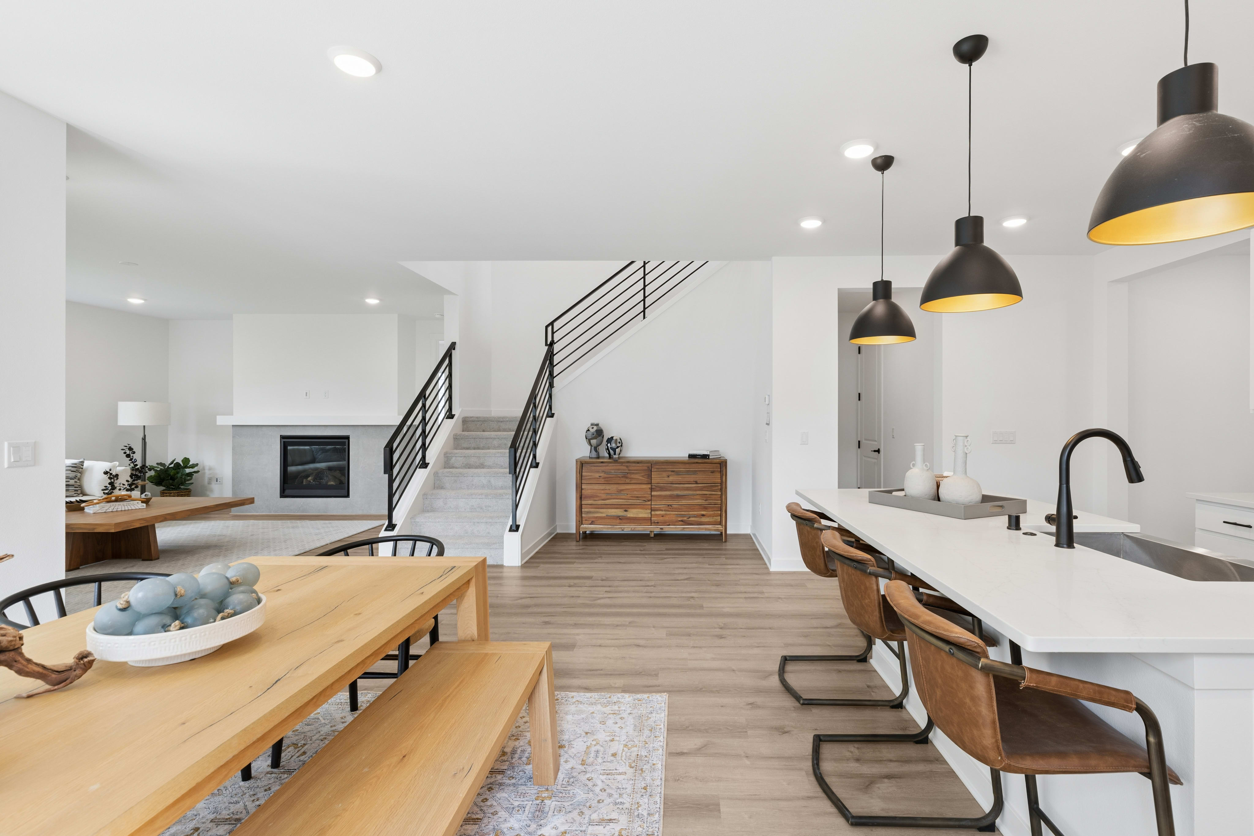 A modern dining area with a wooden table and bench is in the foreground, while a kitchen island with bar stools and a staircase leading upstairs are in the background.