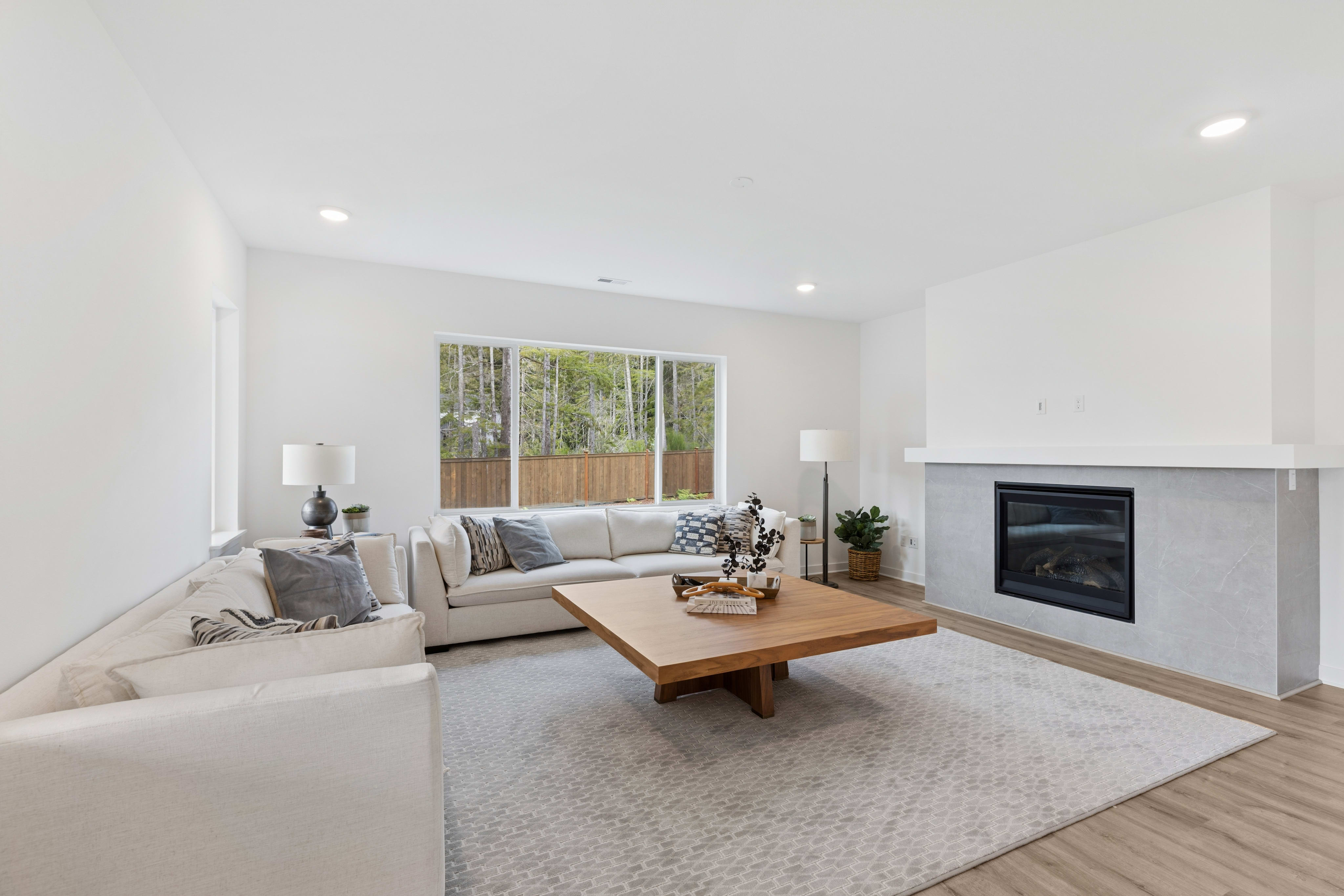 A modern living room with a large white sectional sofa and a wooden coffee table in the foreground, and a fireplace and a large window overlooking trees in the background.