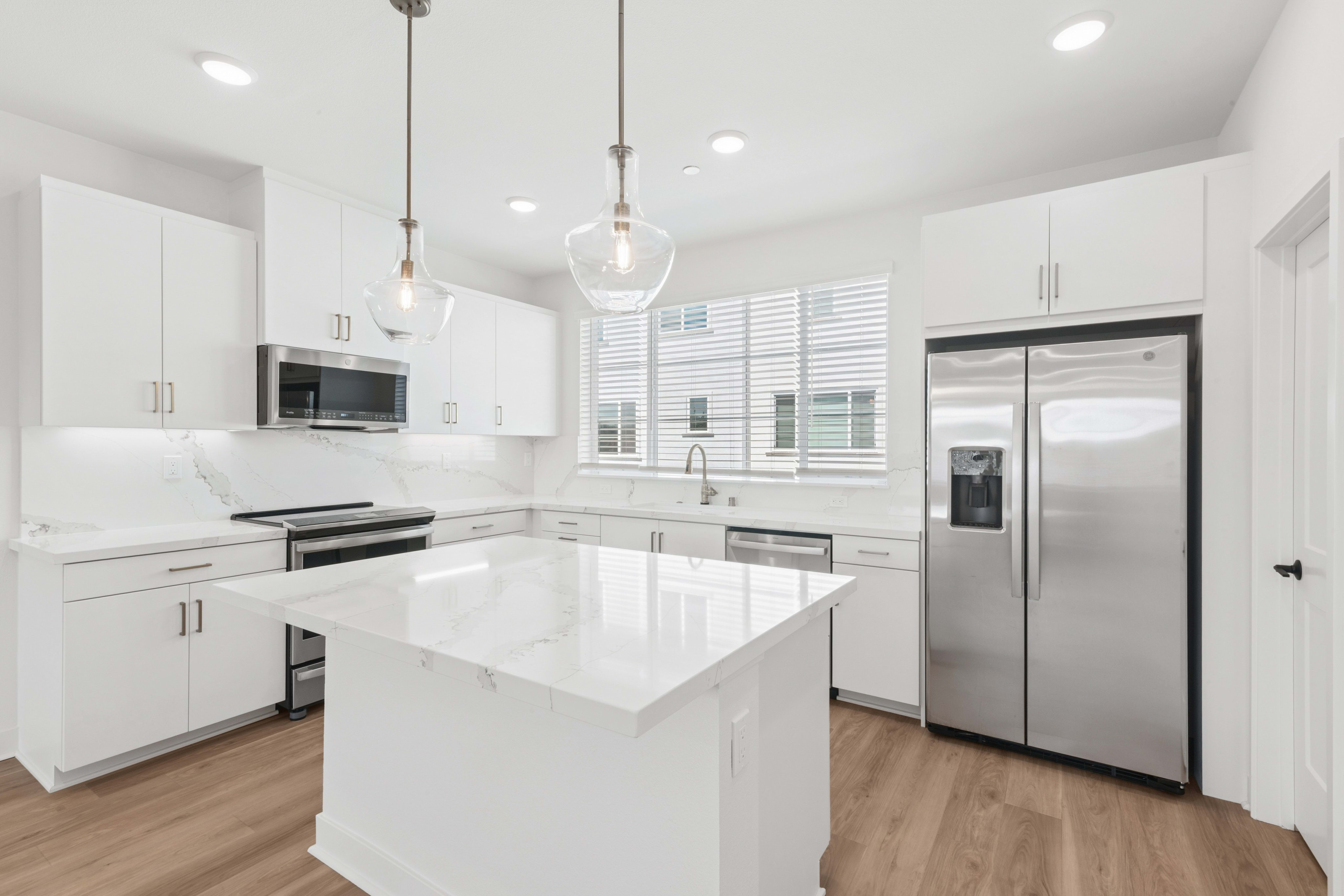 A modern white kitchen with a marble island in the foreground and a window with blinds in the background.