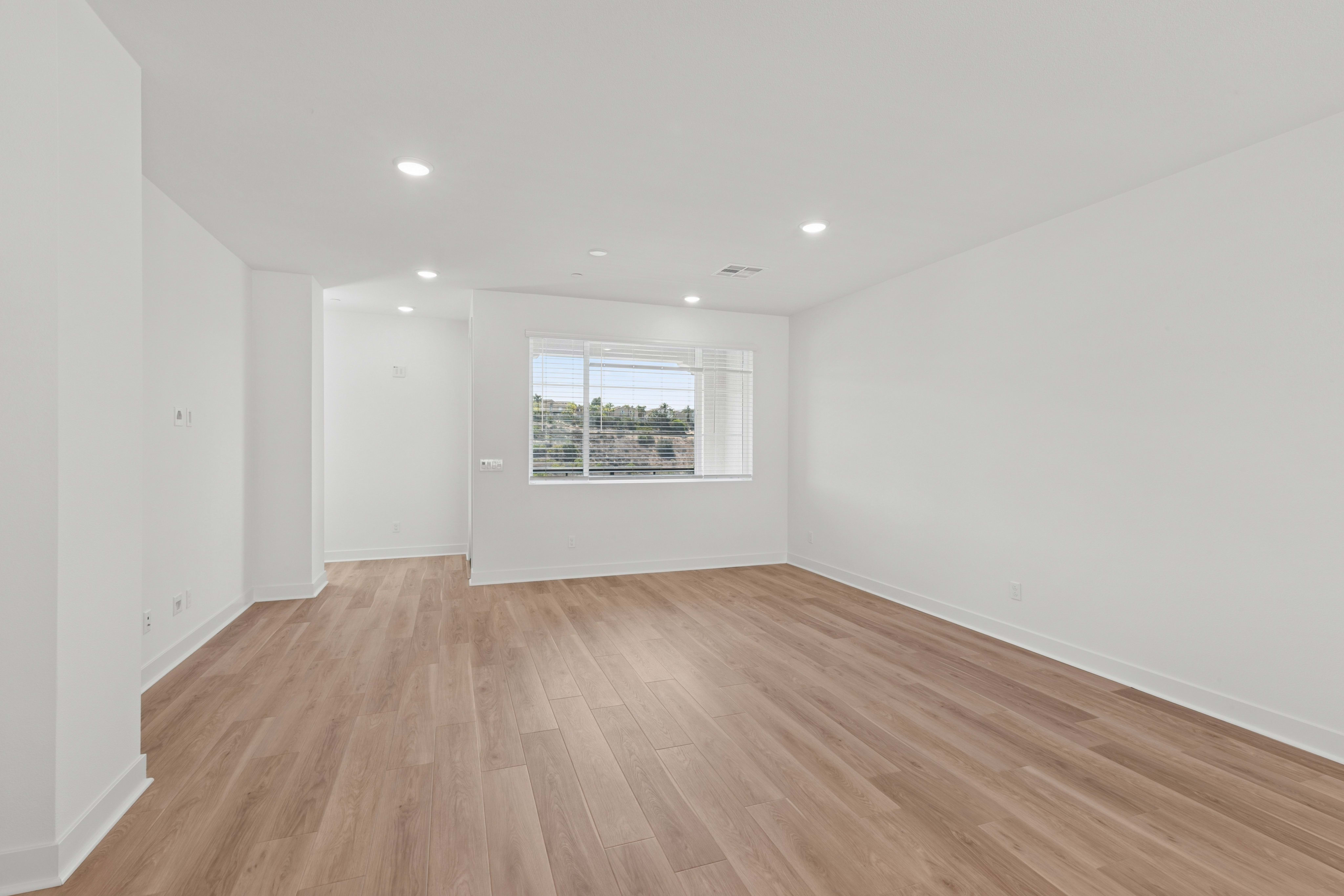 A bright, empty room with light wood flooring in the foreground and a large window revealing a green, hilly landscape in the background.