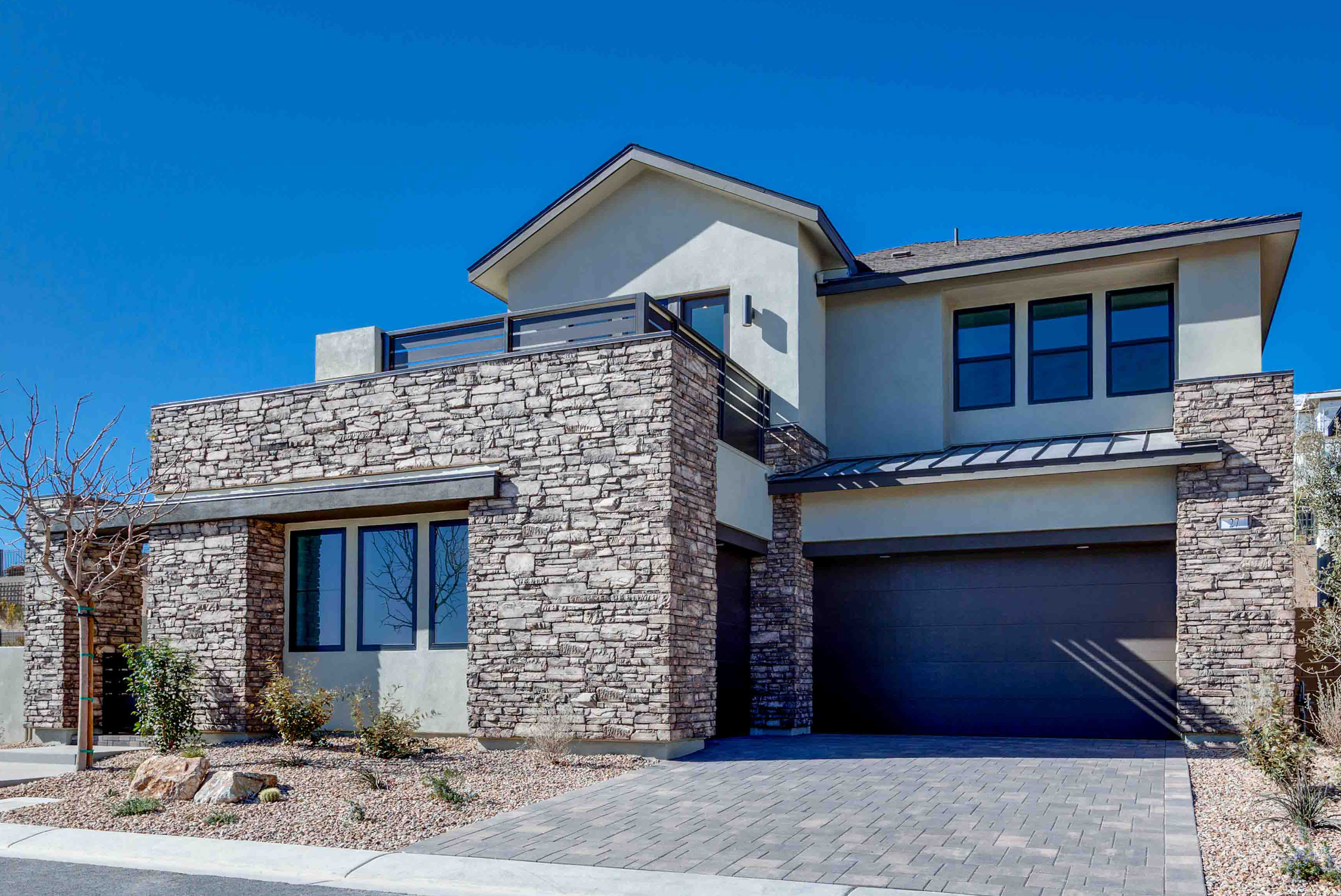 A modern stone-clad house with a dark garage door sits on a paved driveway under a clear blue sky.