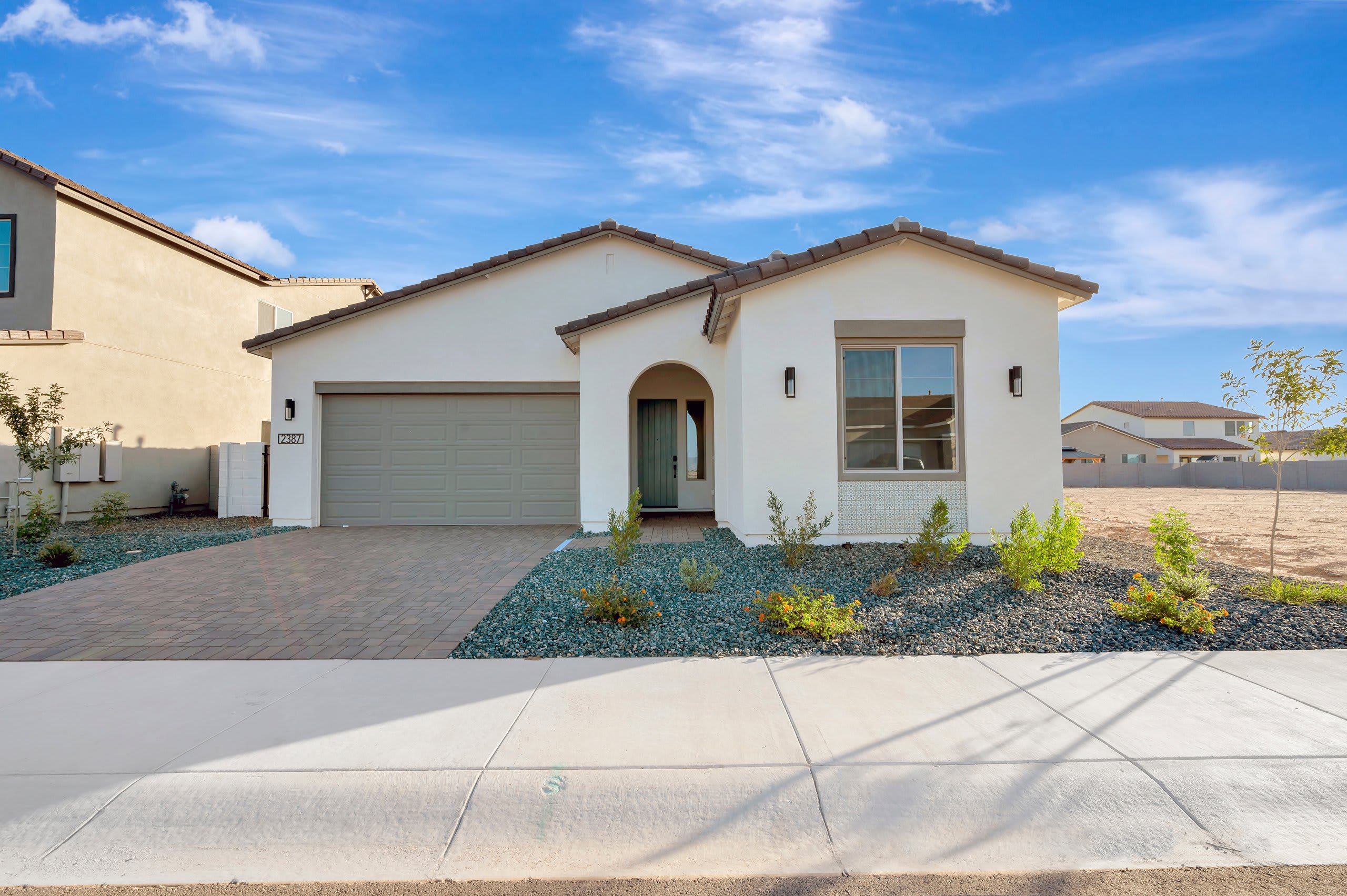 A modern white house with a gray garage door and arched entryway sits behind a sidewalk and a driveway paved with brick, with other houses and a bright blue sky in the background.