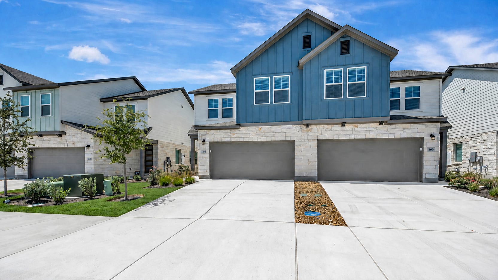A modern townhouse with a blue facade and stone accents sits behind a concrete driveway, with a clear blue sky and scattered clouds in the background.