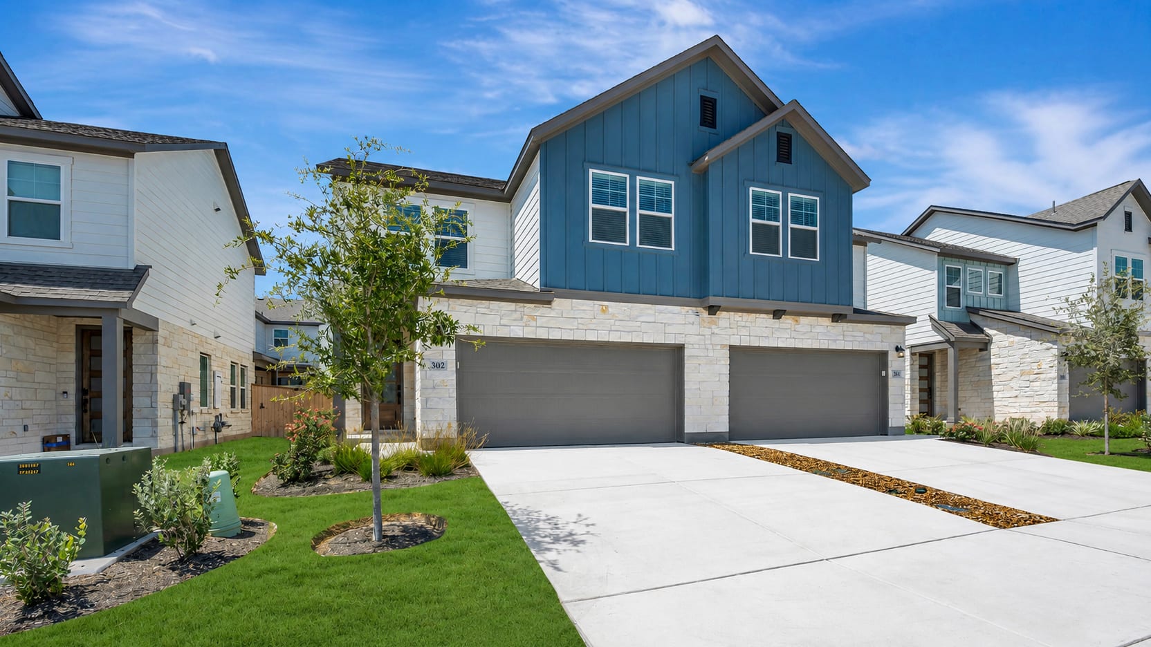 A modern townhouse with blue siding and stone accents features a double garage in the foreground, while other similar townhouses line the street under a bright blue sky in the background.