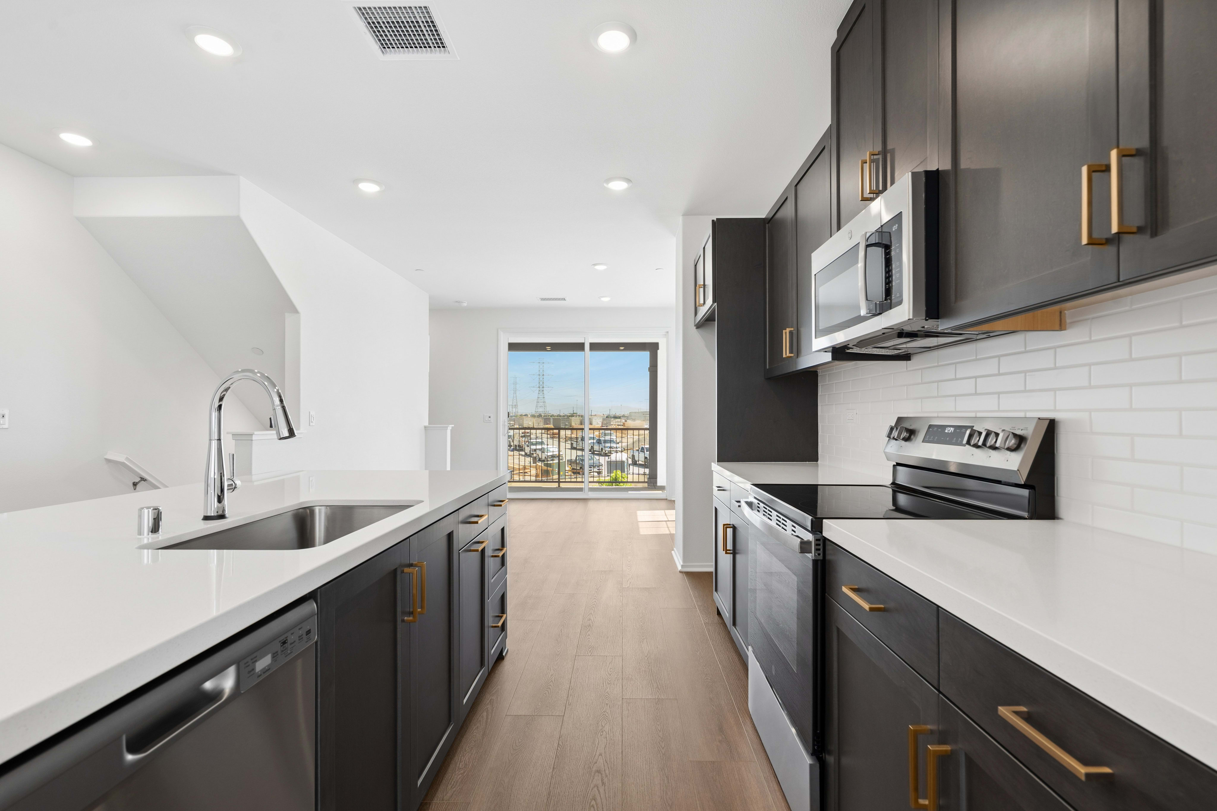 A modern kitchen with dark cabinets and white countertops in the foreground opens to a living area with a view of a marina in the background.