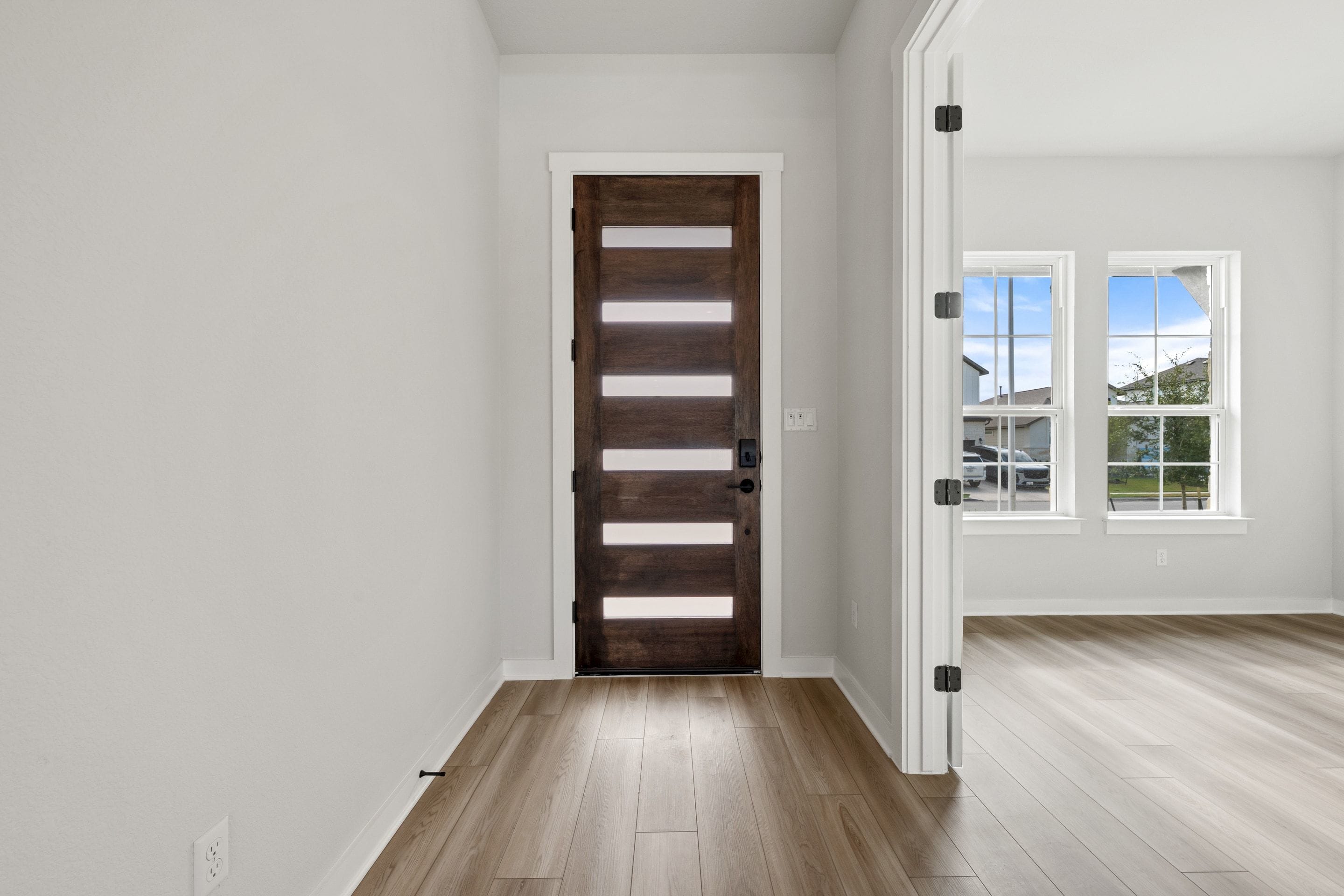 A modern wooden door with horizontal glass panels stands in the foreground, leading into a bright room with large windows in the background.