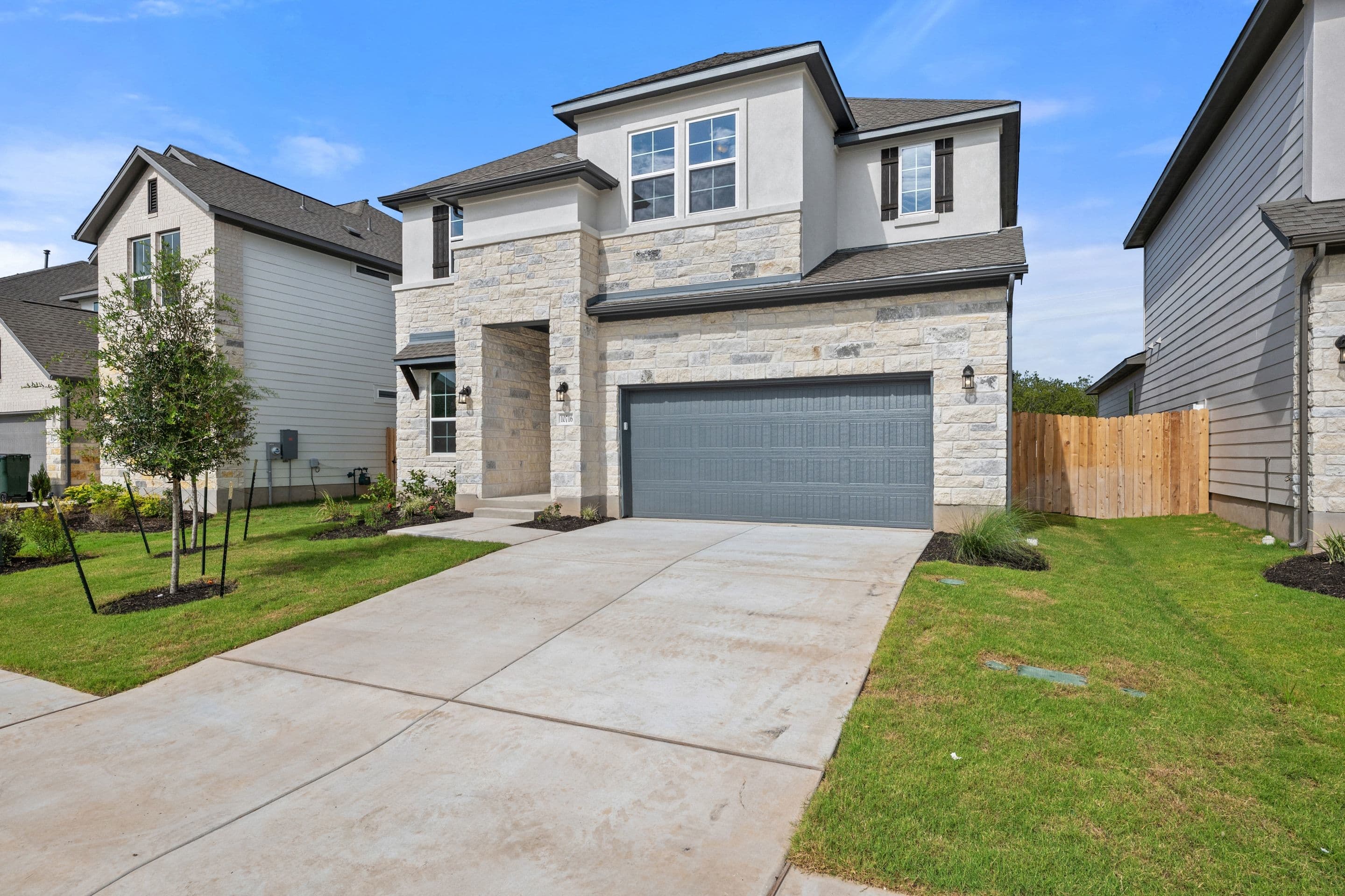 A modern stone-clad house with a gray garage door sits on a concrete driveway in the foreground, while neighboring houses and a wooden fence are visible in the background under a blue sky.