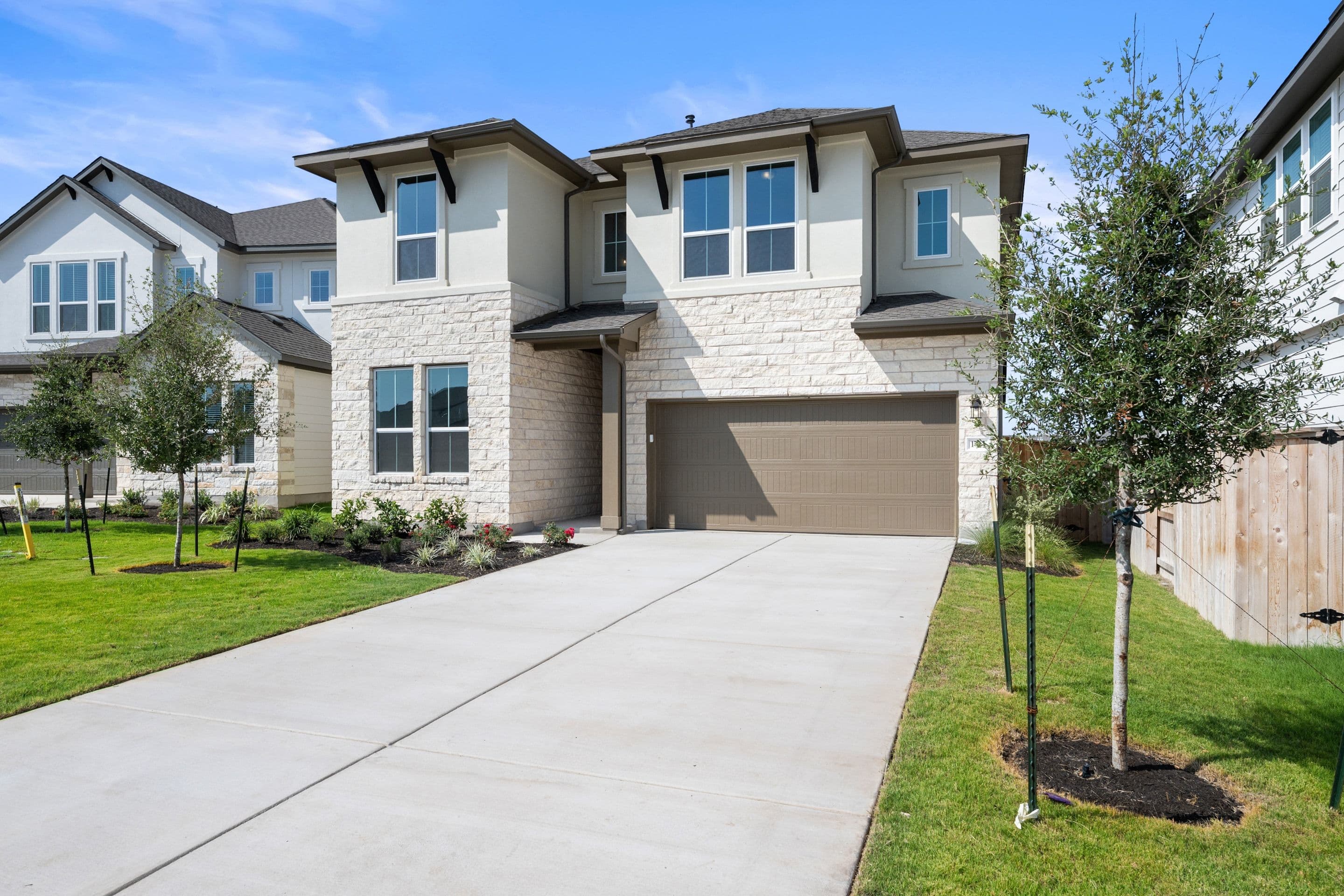 A modern two-story house with a stone facade and a beige garage door sits on a manicured lawn, with other houses and a clear blue sky in the background.