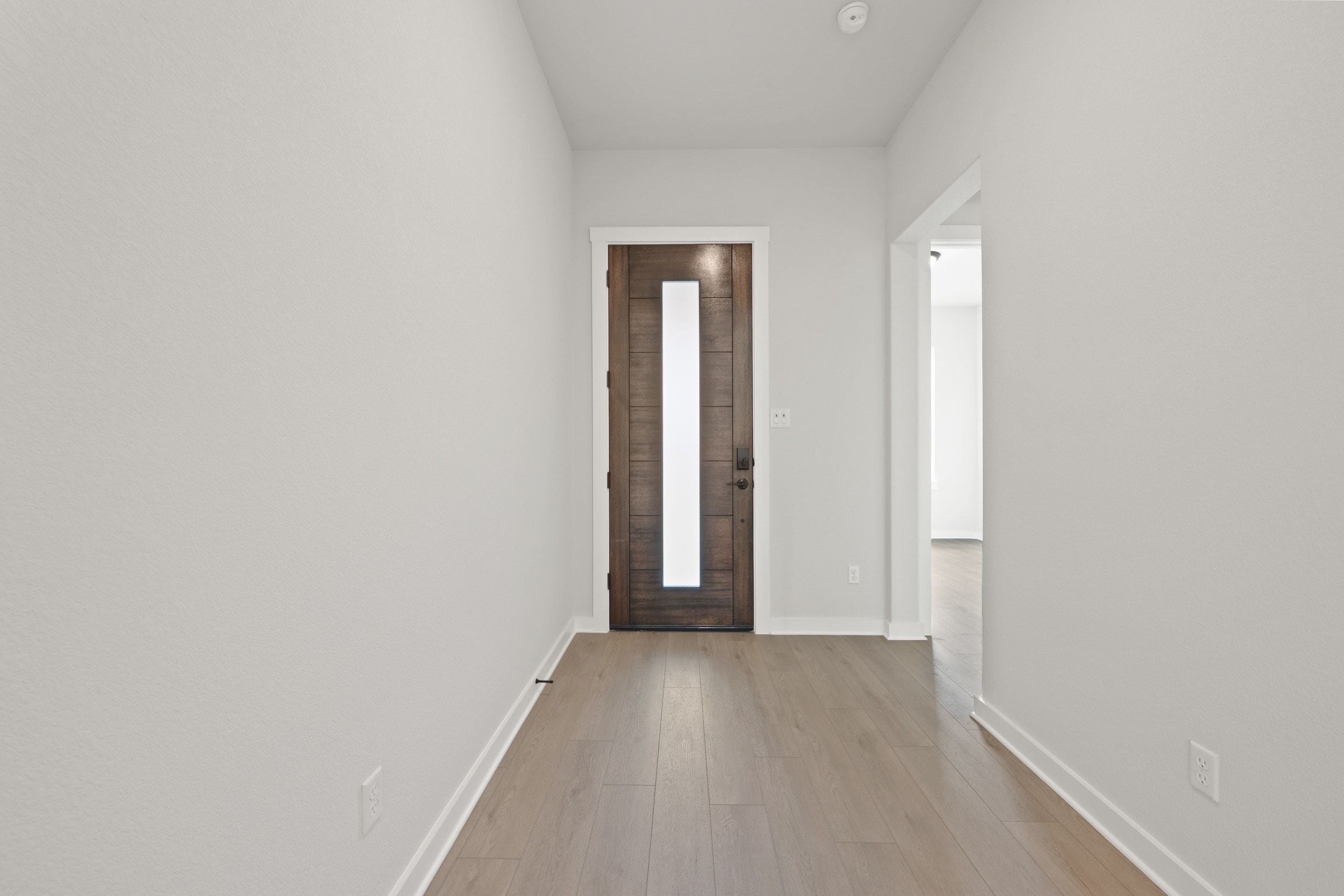 A modern hallway with a dark wood door and a light wood floor leads to a brightly lit room in the background.