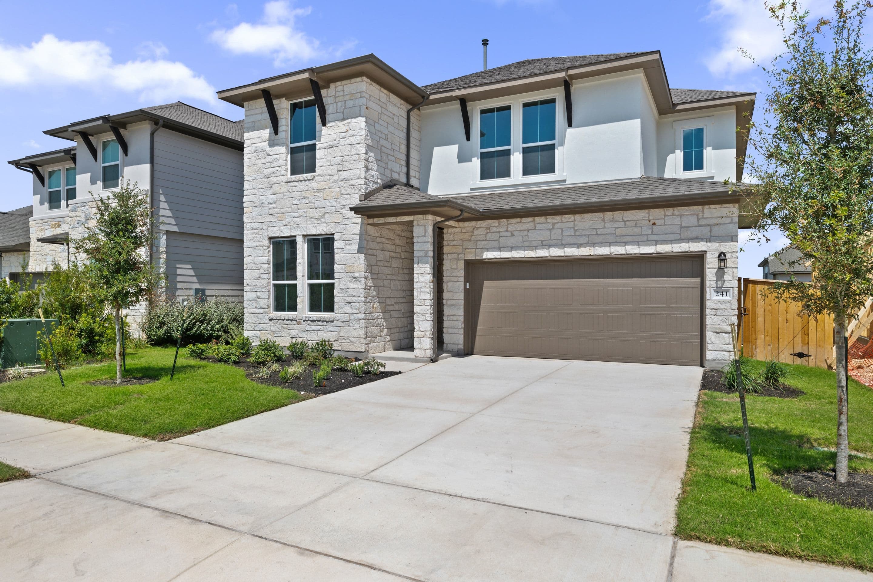 A modern two-story house with white stone accents and a brown garage door sits on a concrete driveway, with a manicured lawn in the foreground and other houses and trees in the background under a blue sky.