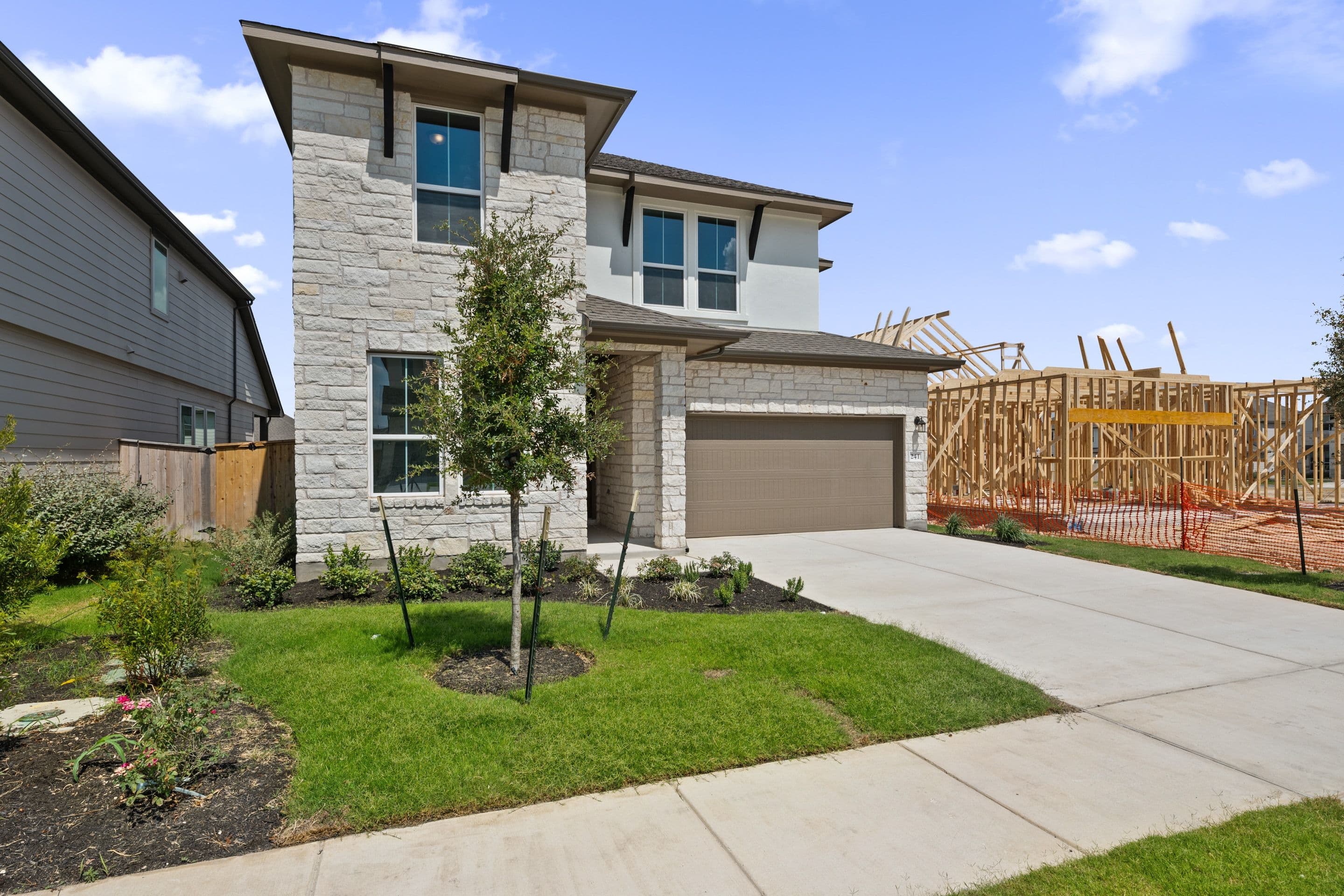 A modern stone-clad house with a manicured lawn in the foreground stands before a construction site with wooden framing in the background.