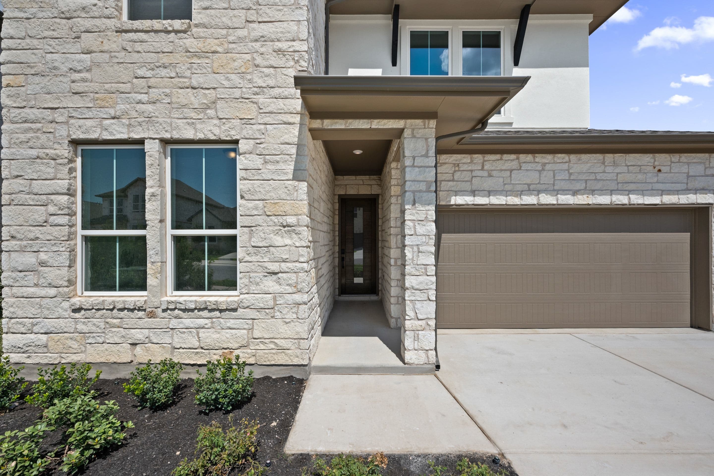A modern stone-clad house with a light brown garage door and a walkway leading to the front door is set against a bright sky.